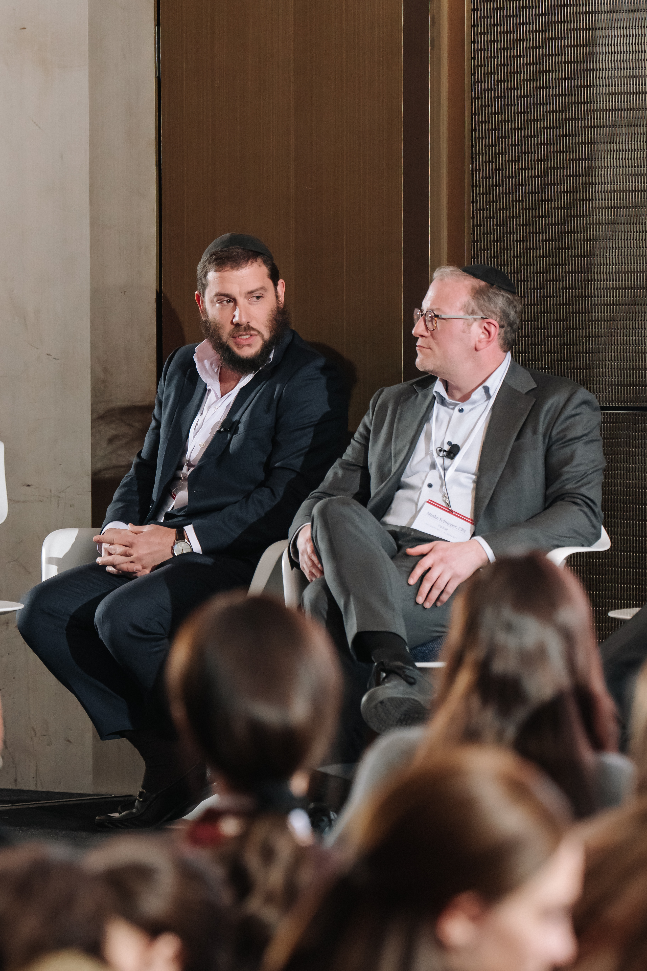 Two men in formal attire seated on a panel at an indoor event, with an audience visible in the foreground.