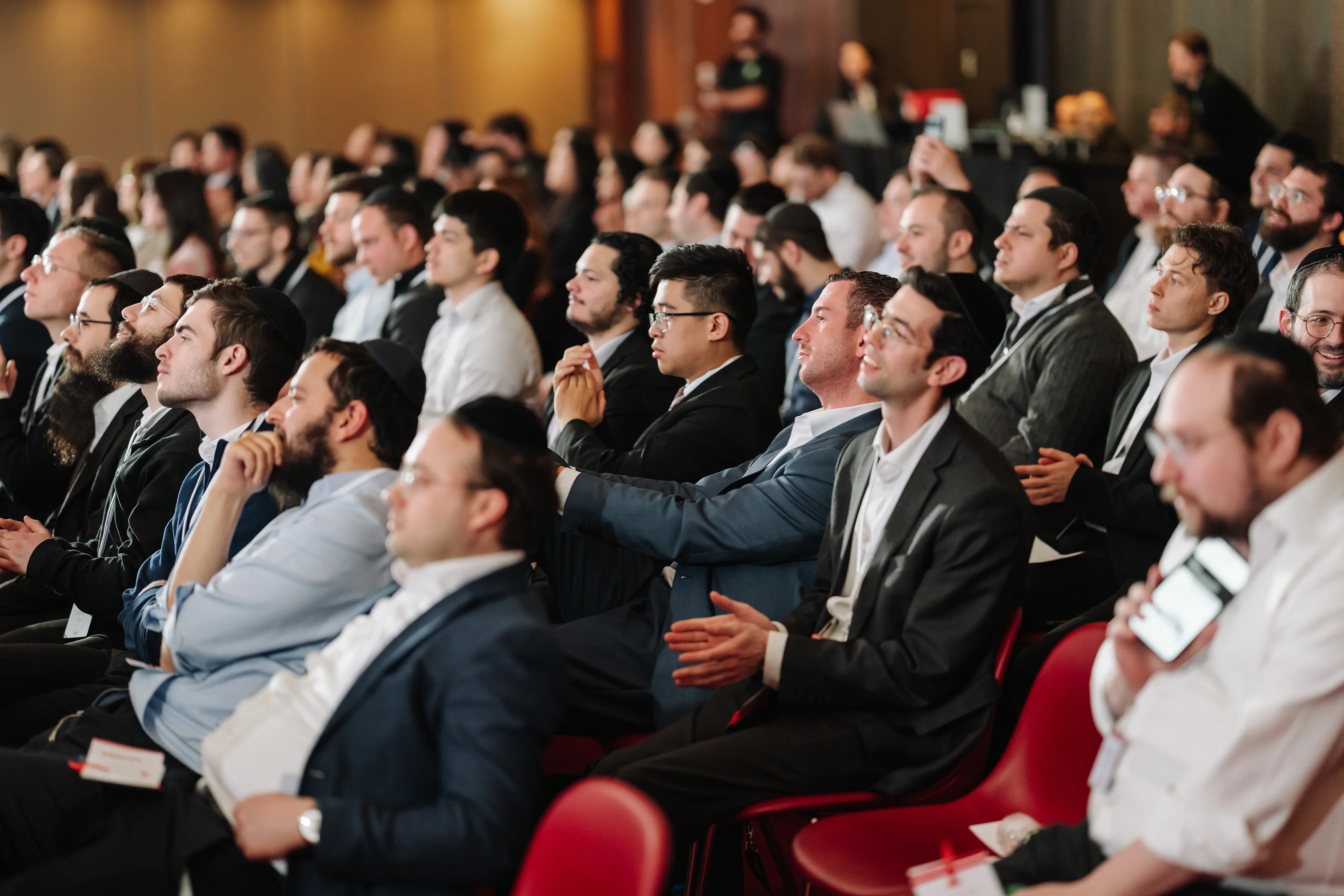Wedding guests seated in a ceremony hall during a formal moment, seen from the side in warm indoor lighting.