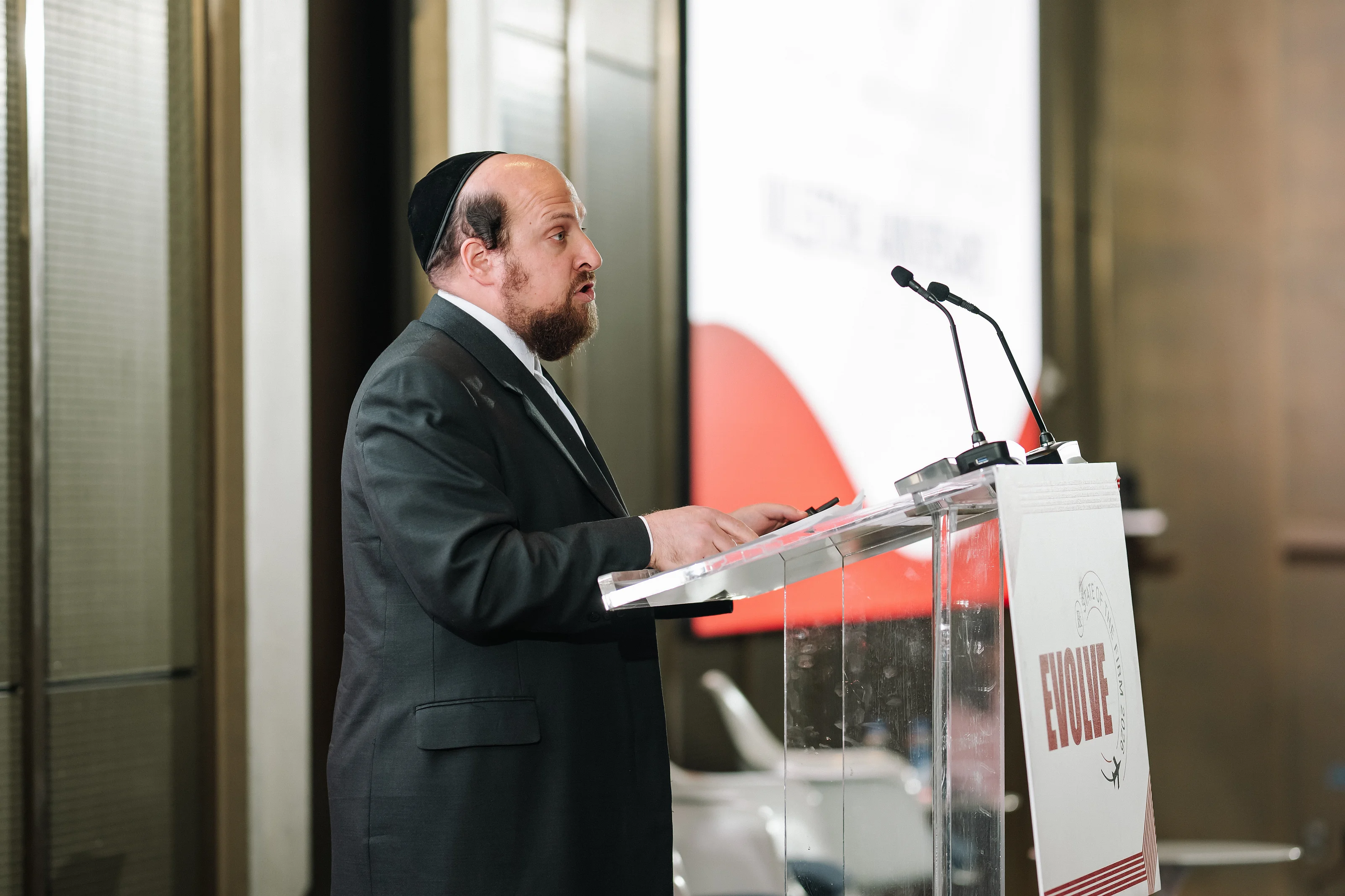 Man in a suit speaking at a podium with microphones during a Jewish celebration, viewed in side profile.