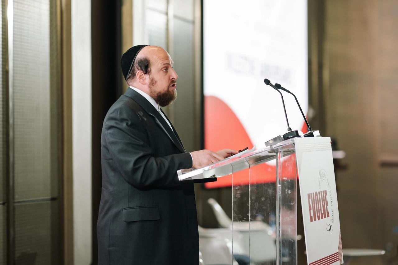 Man in a suit speaking at a podium with microphones during a Jewish celebration, viewed in side profile.