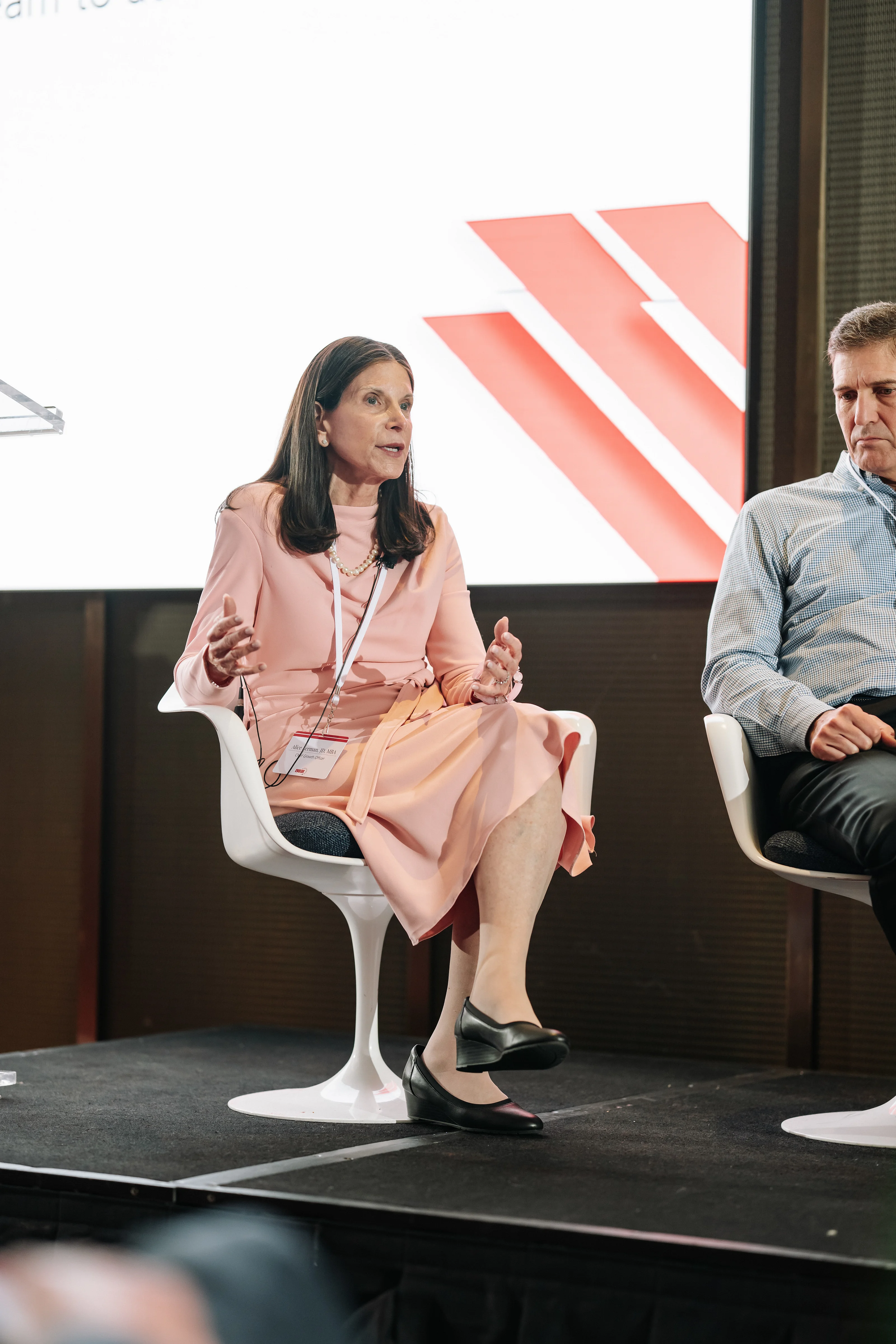 Woman in a light pink outfit seated on a stage in front of a screen with red-and-white diagonal stripes, gesturing while speaking.