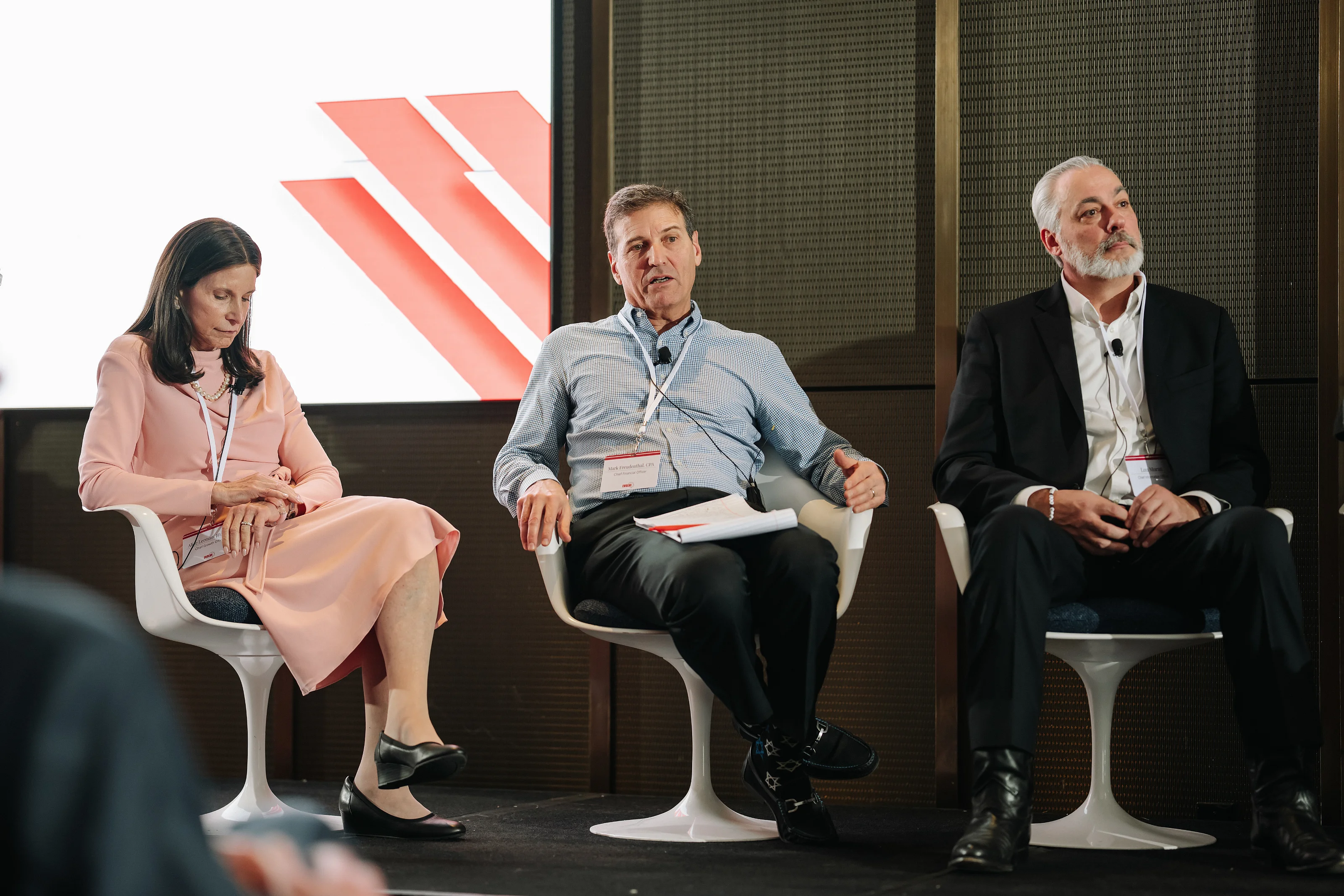 Three panel speakers seated on stage with microphones and papers in front of a projection screen.