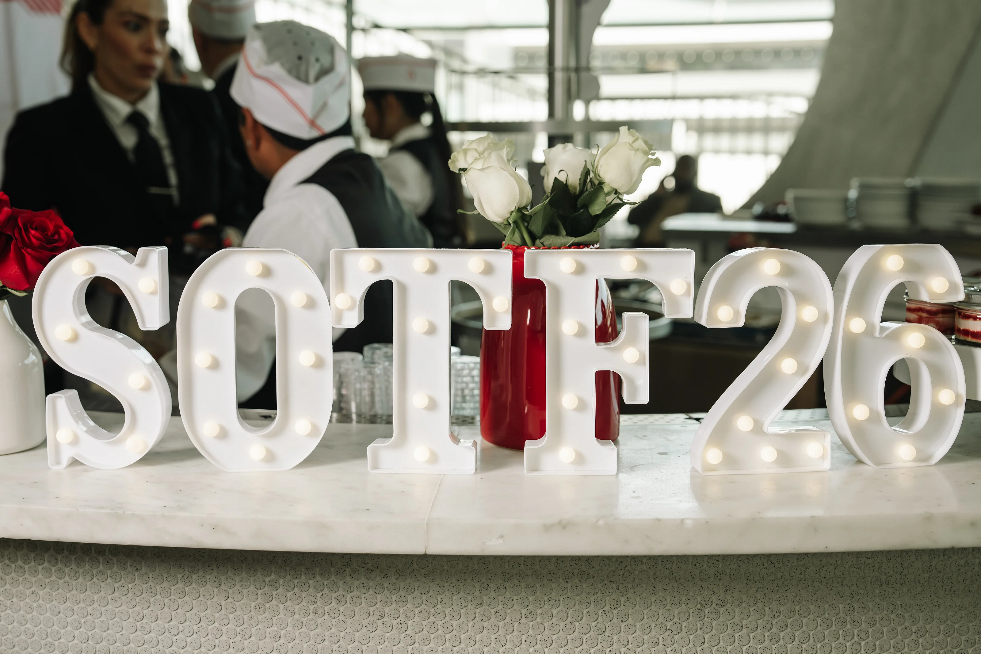 Lit white marquee letters spelling “SOFTLY” with flowers on a celebration table, guests blurred in the background.