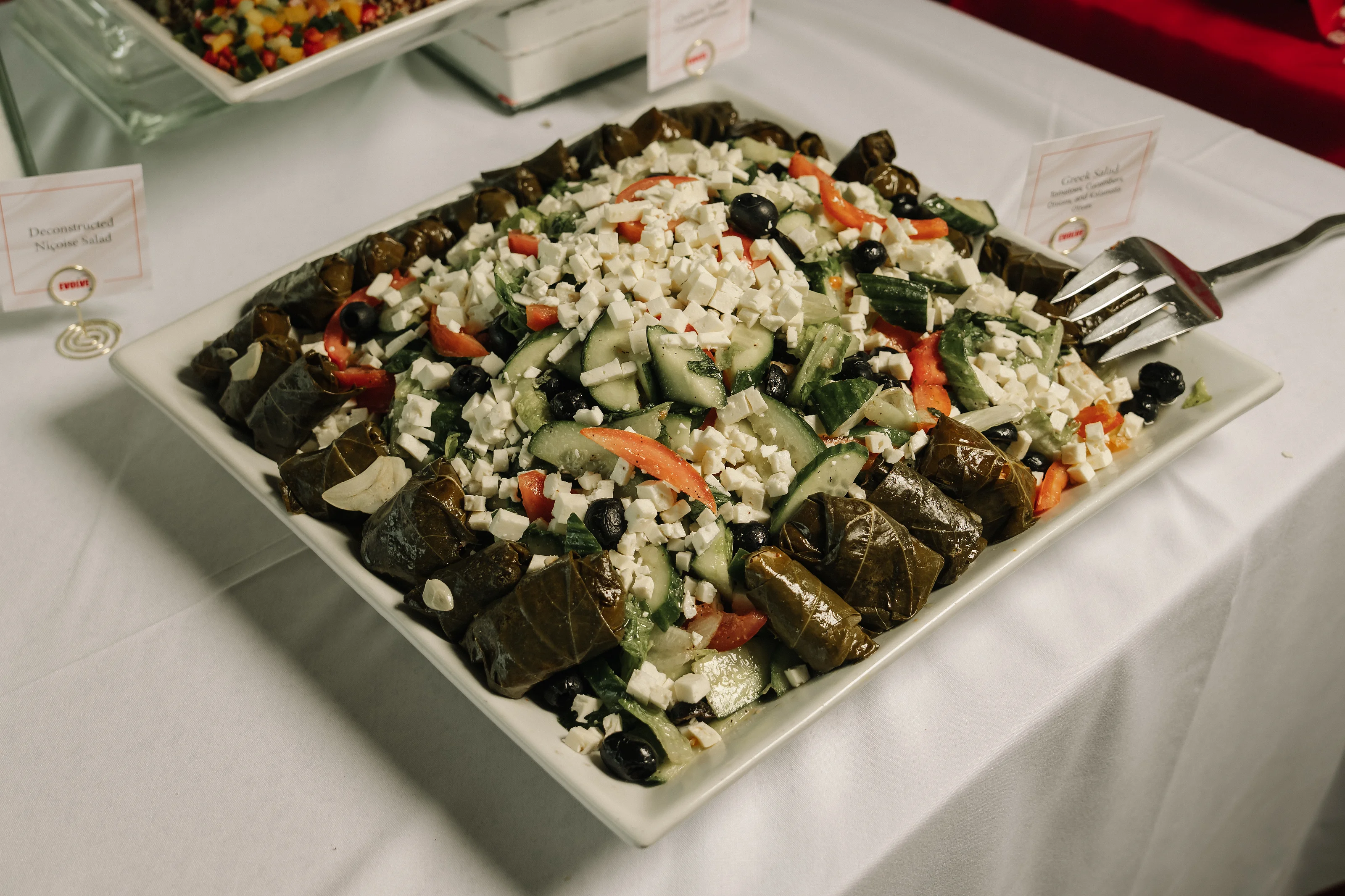 Overhead view of a rectangular serving platter with stuffed grape leaves and chopped salad on a white tablecloth.