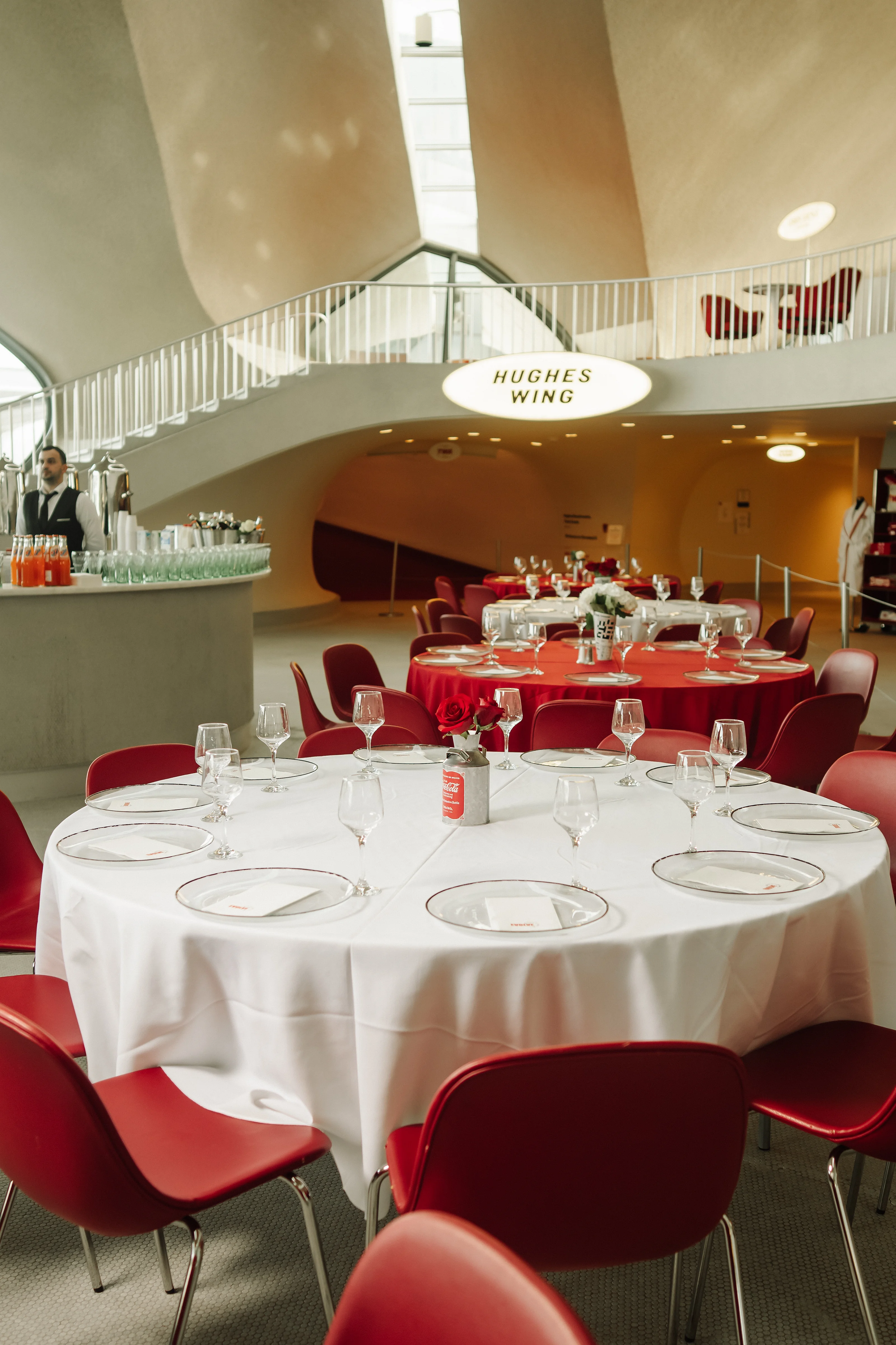 Reception table with white linens, red chairs, and glassware set for a wedding ceremony in a curved indoor venue.