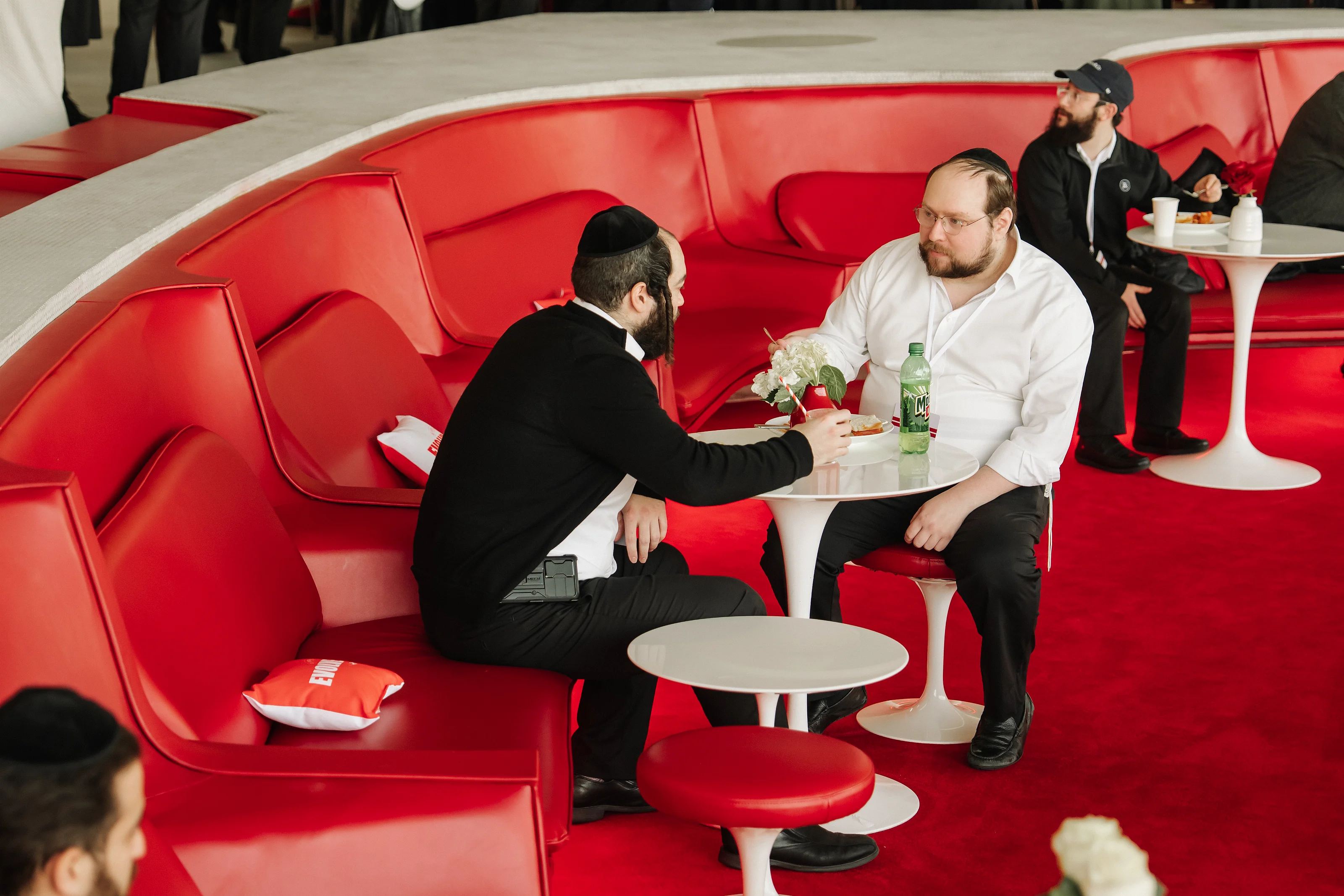 Two seated men at a round table with flowers on a small table in front of bright red cushioned seating during an event.