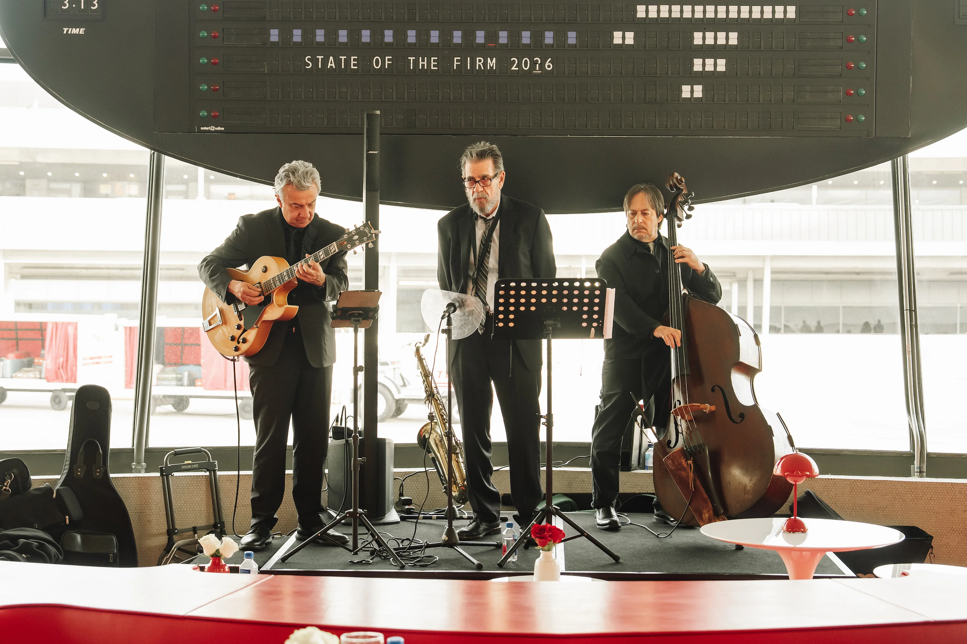 Wedding reception stage with three musicians performing string and guitar, microphones and music stands visible, backlit by bright windows.