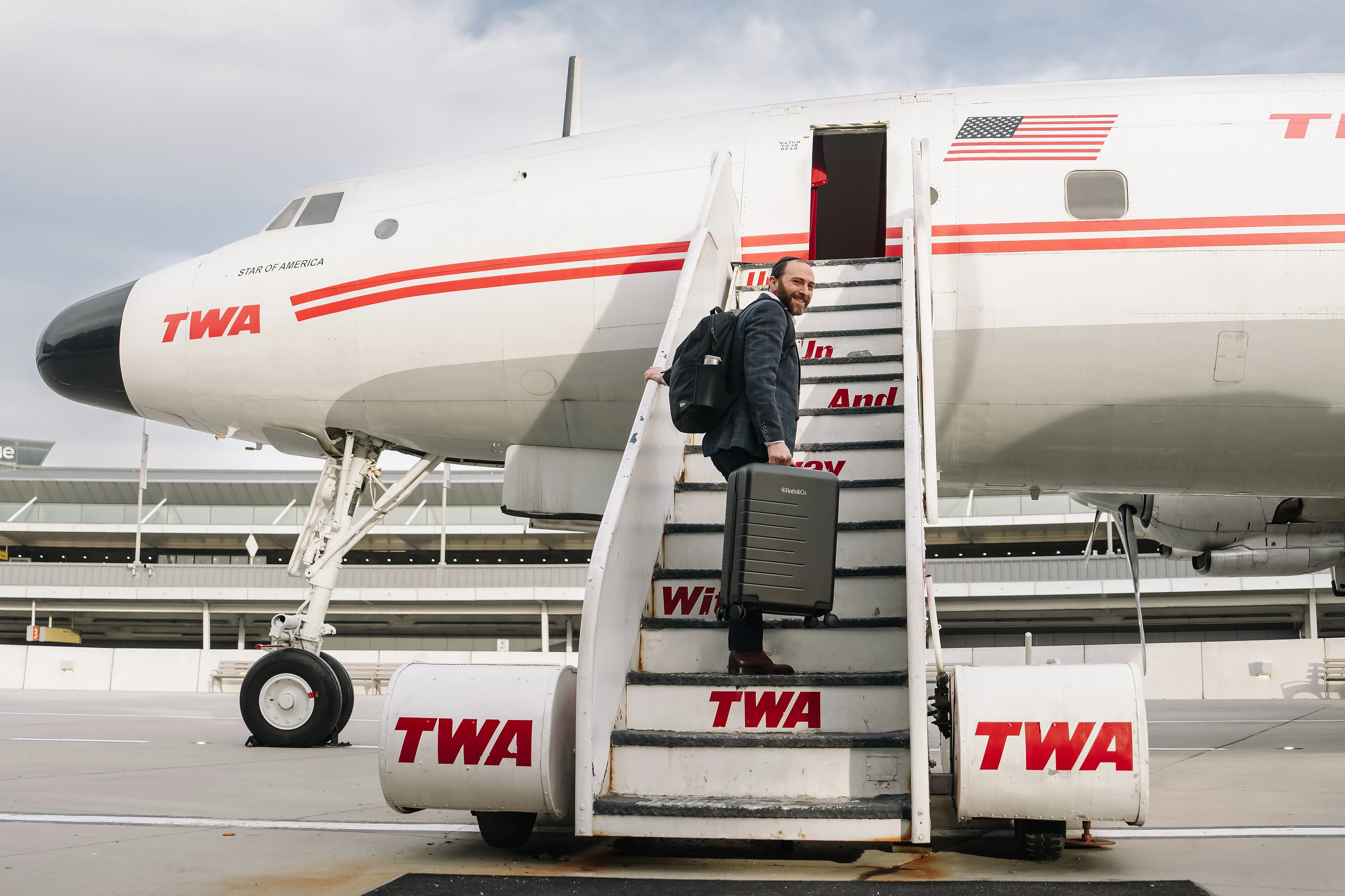 Man descending airplane stairs with a suitcase in an airport, cloudy sky and large aircraft in frame.