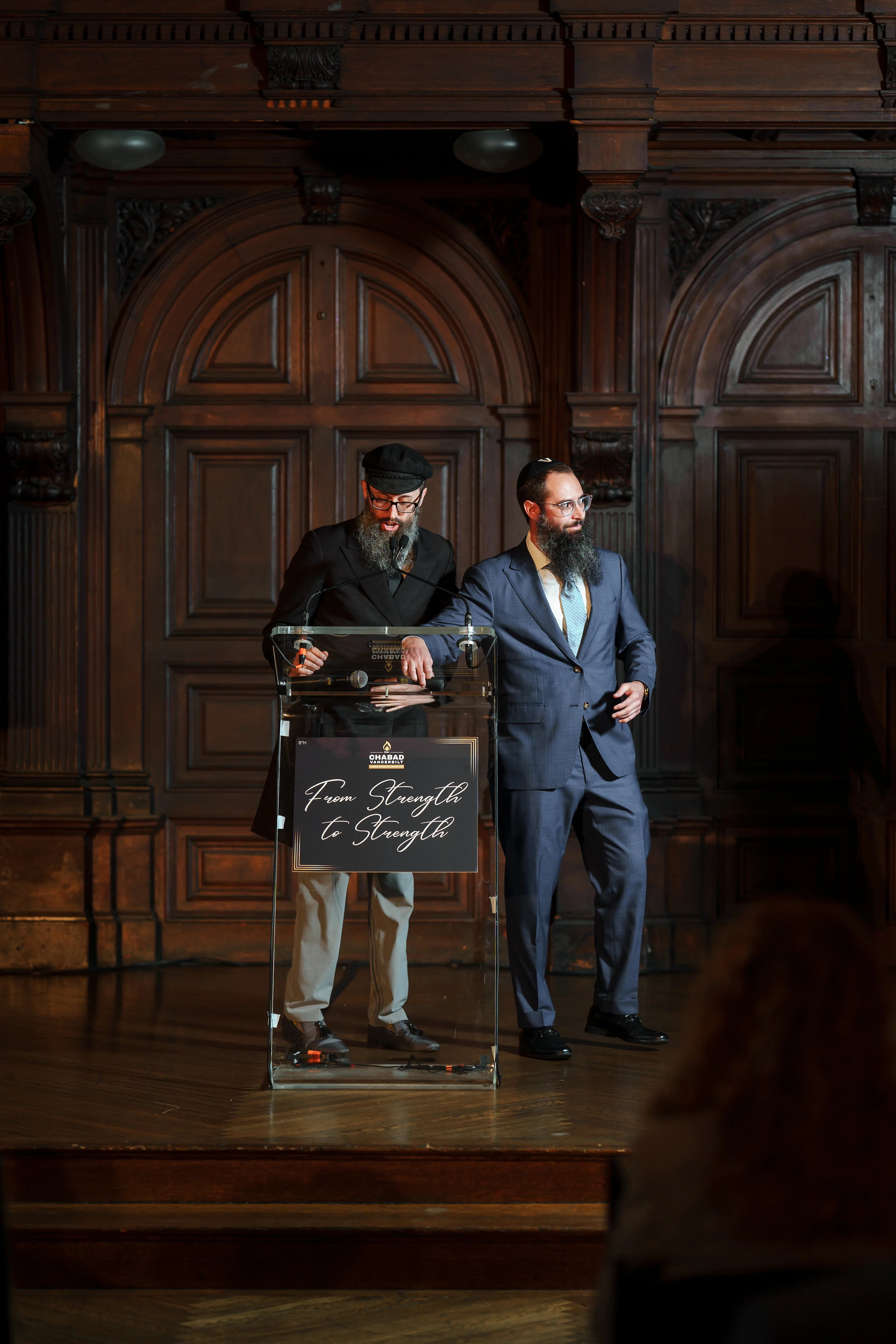 Two men in formal attire with yarmulkes during a ritual moment at a podium in a warmly lit, photographed by CJ Studios