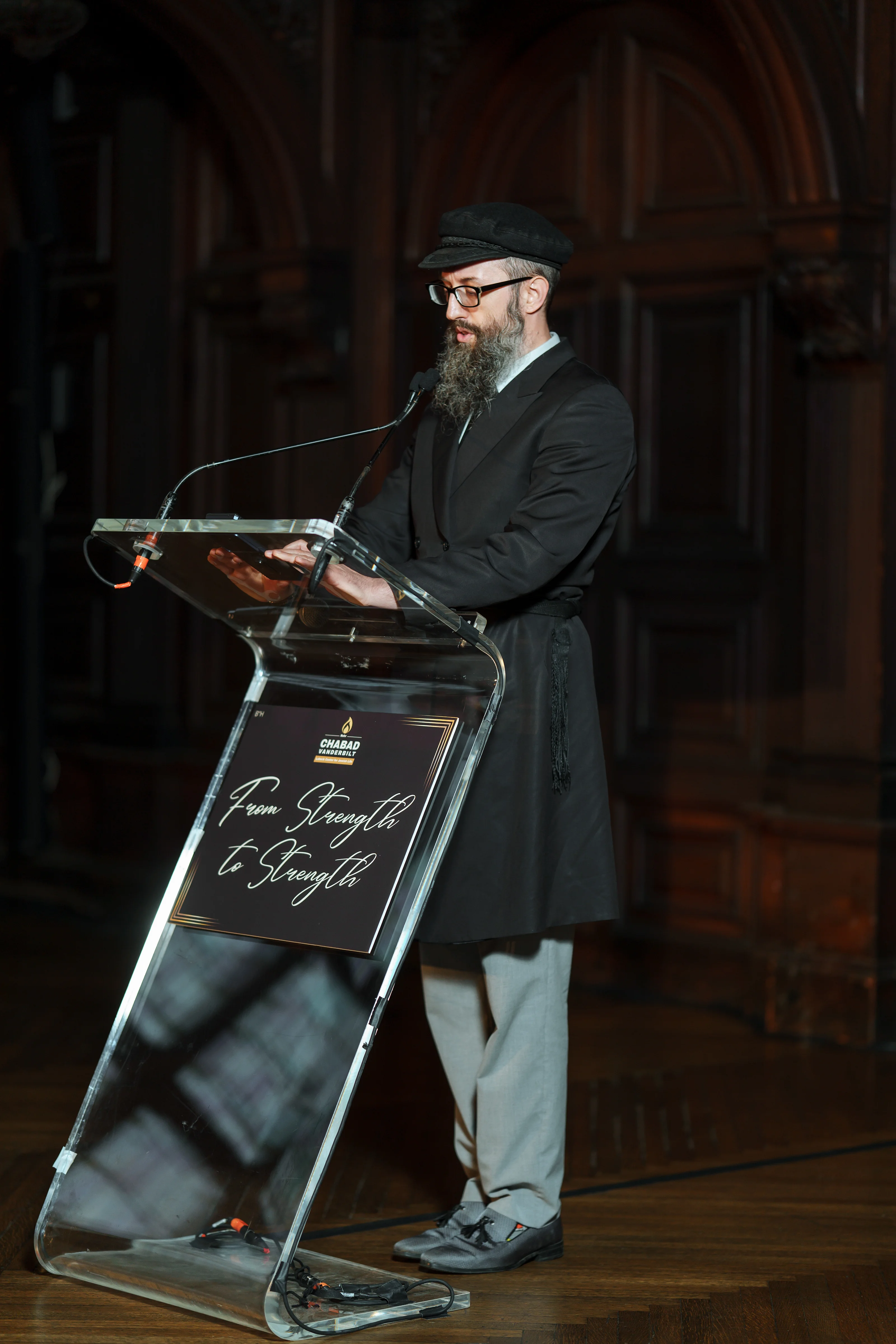 Framed photo of a bearded speaker wearing a kippah and glasses, standing at a podium with a microphone under warm indoor lighting.
