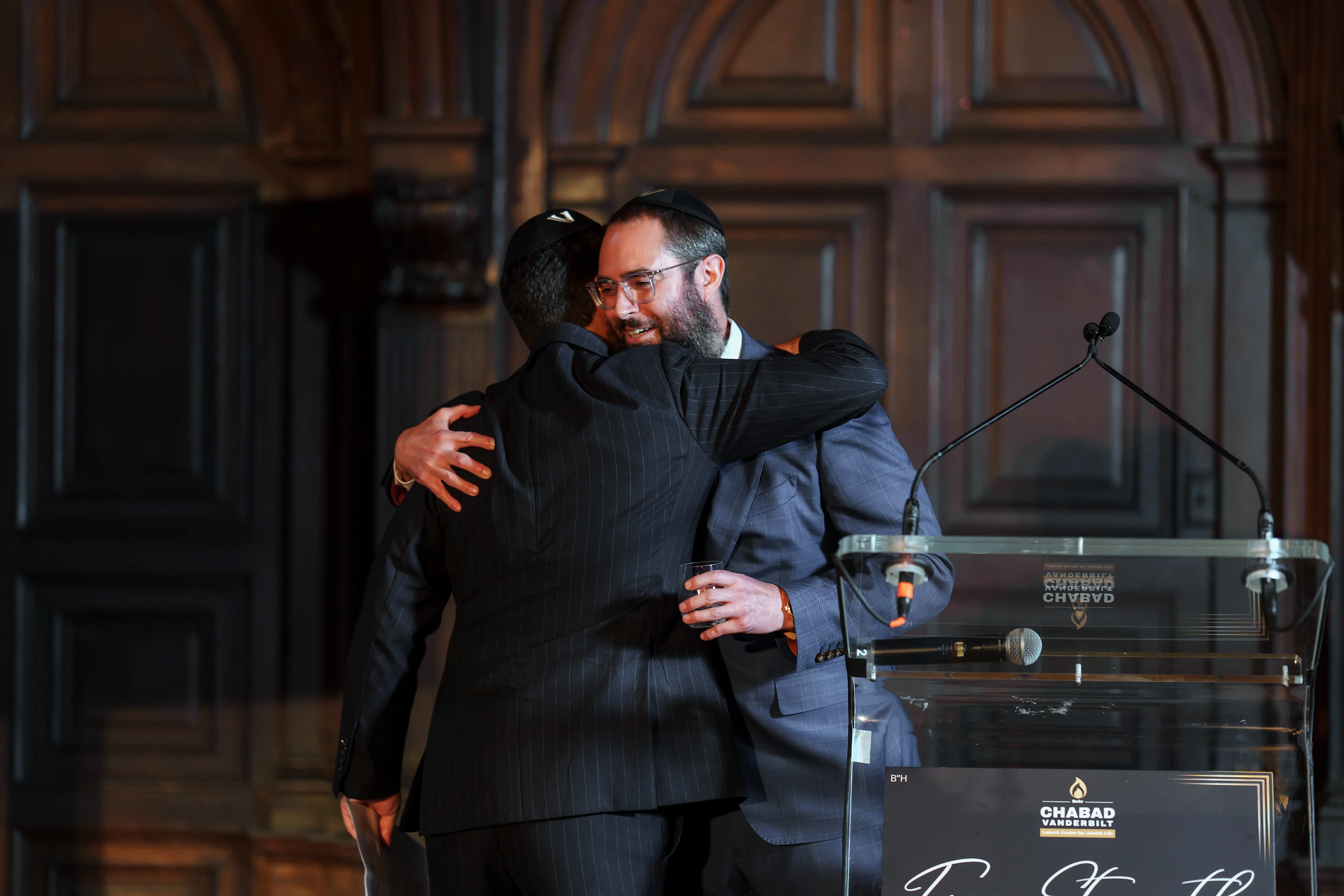 Smiling couple in formal attire sharing a warm hug during a celebratory moment at an event with a microphone podium in the background.