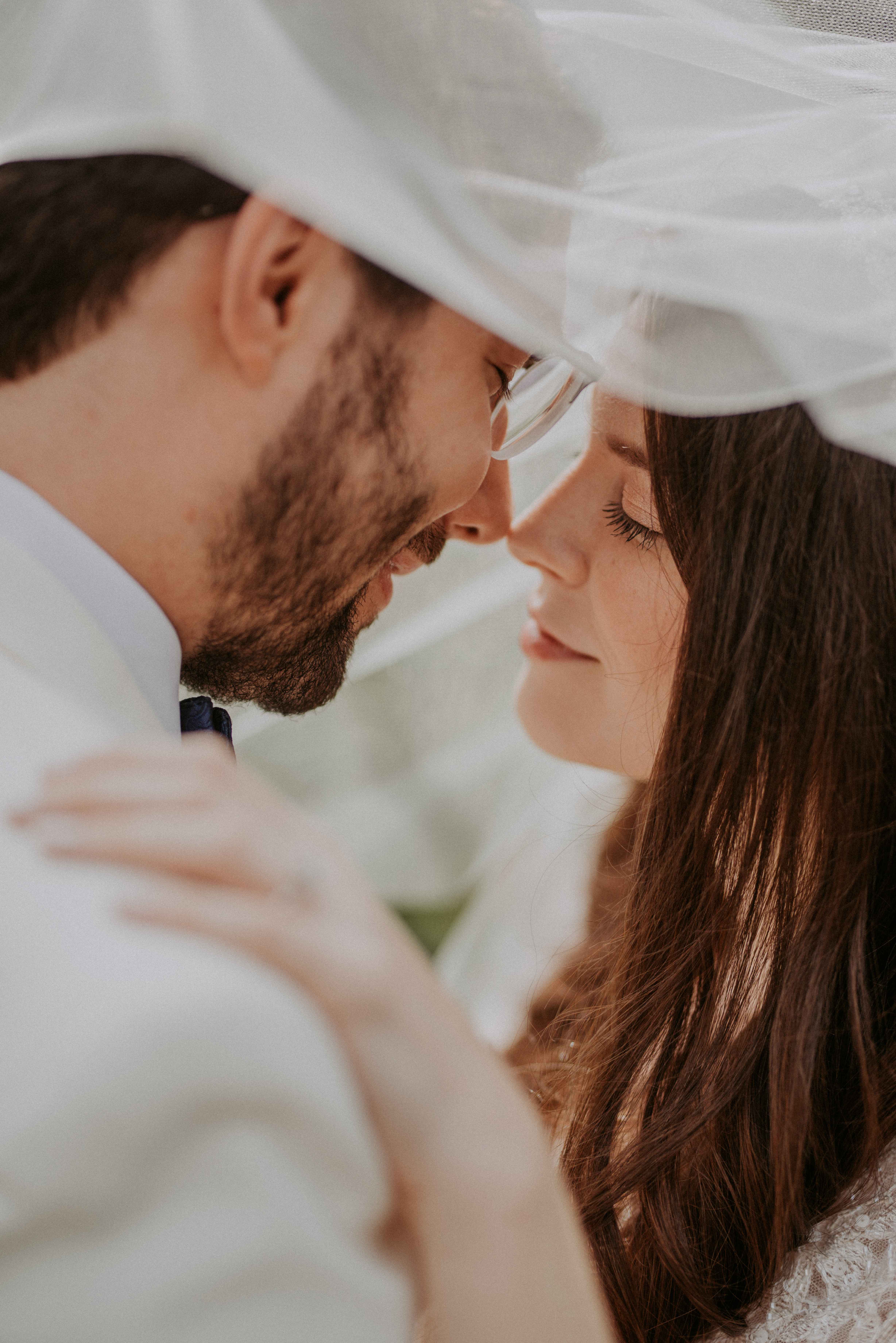 A bride and groom under a white chuppah veil share an intimate moment with eyes closed, foreheads close together.