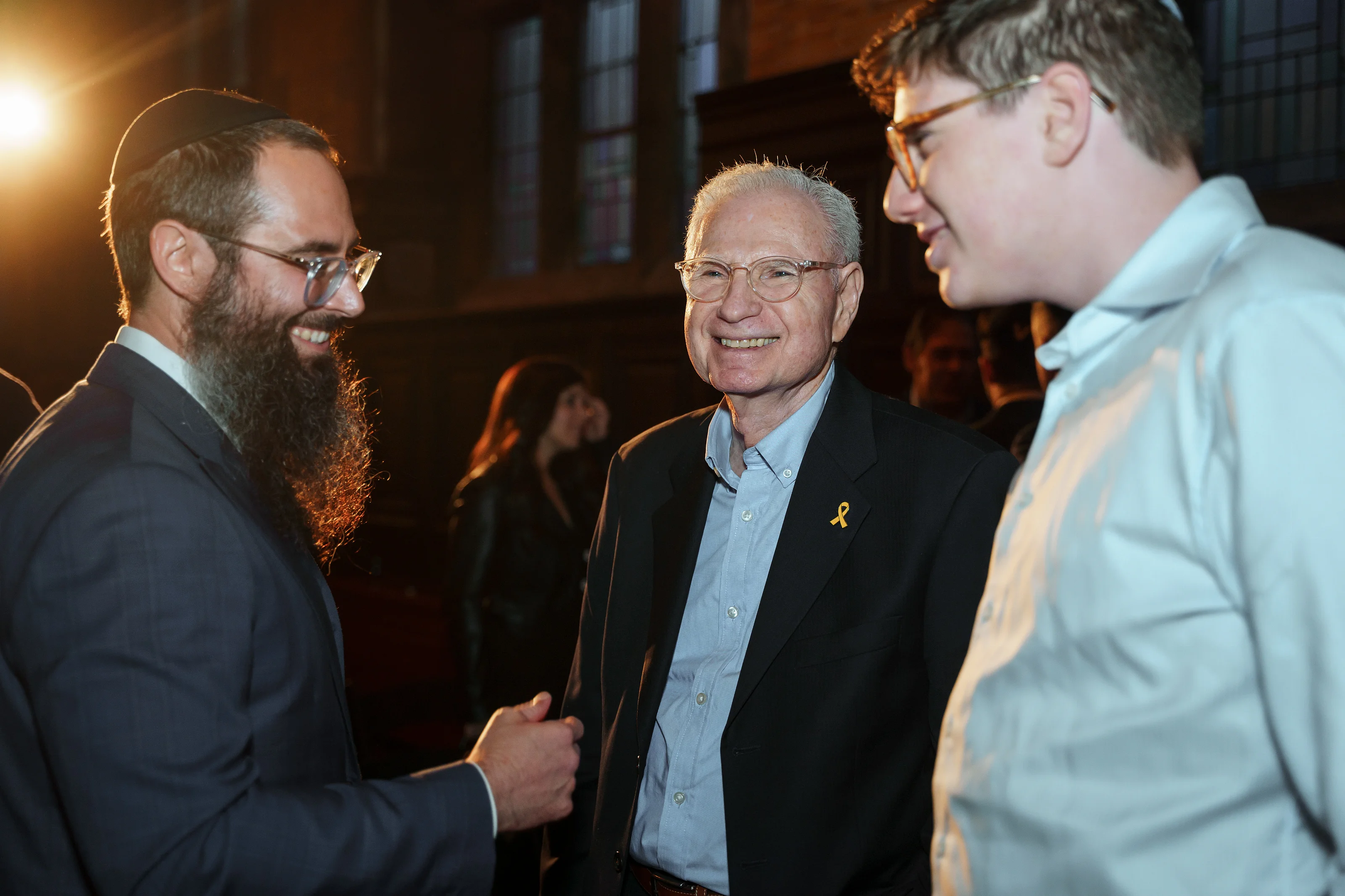 Candid moment of three men smiling and chatting at a Jewish celebration with warm evening lighting in an indoor hall.