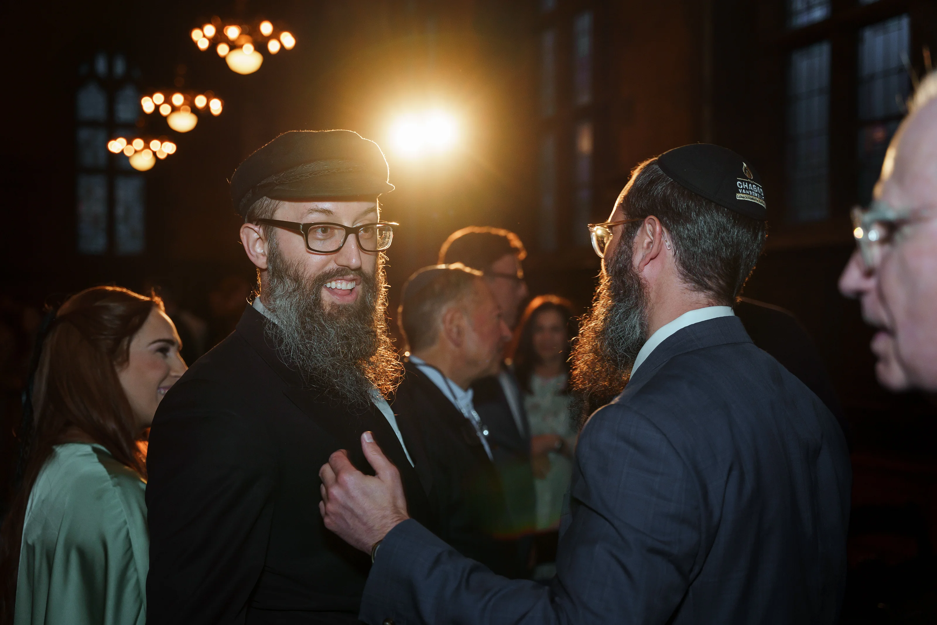 Two bearded men in formal attire share a close embrace and warm smiles during a celebration under warm amber lights.