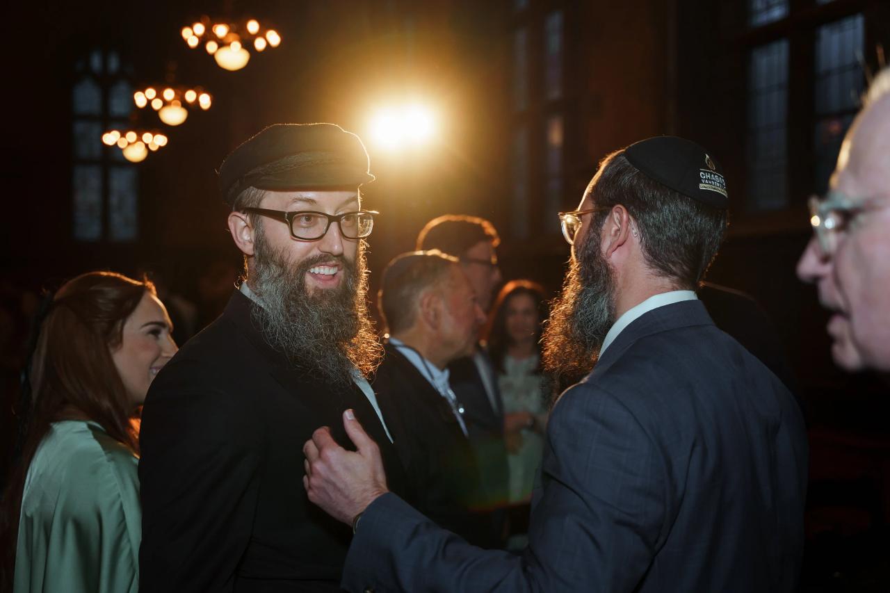 Two bearded men in formal attire share a close embrace and warm smiles during a celebration under warm amber lights.