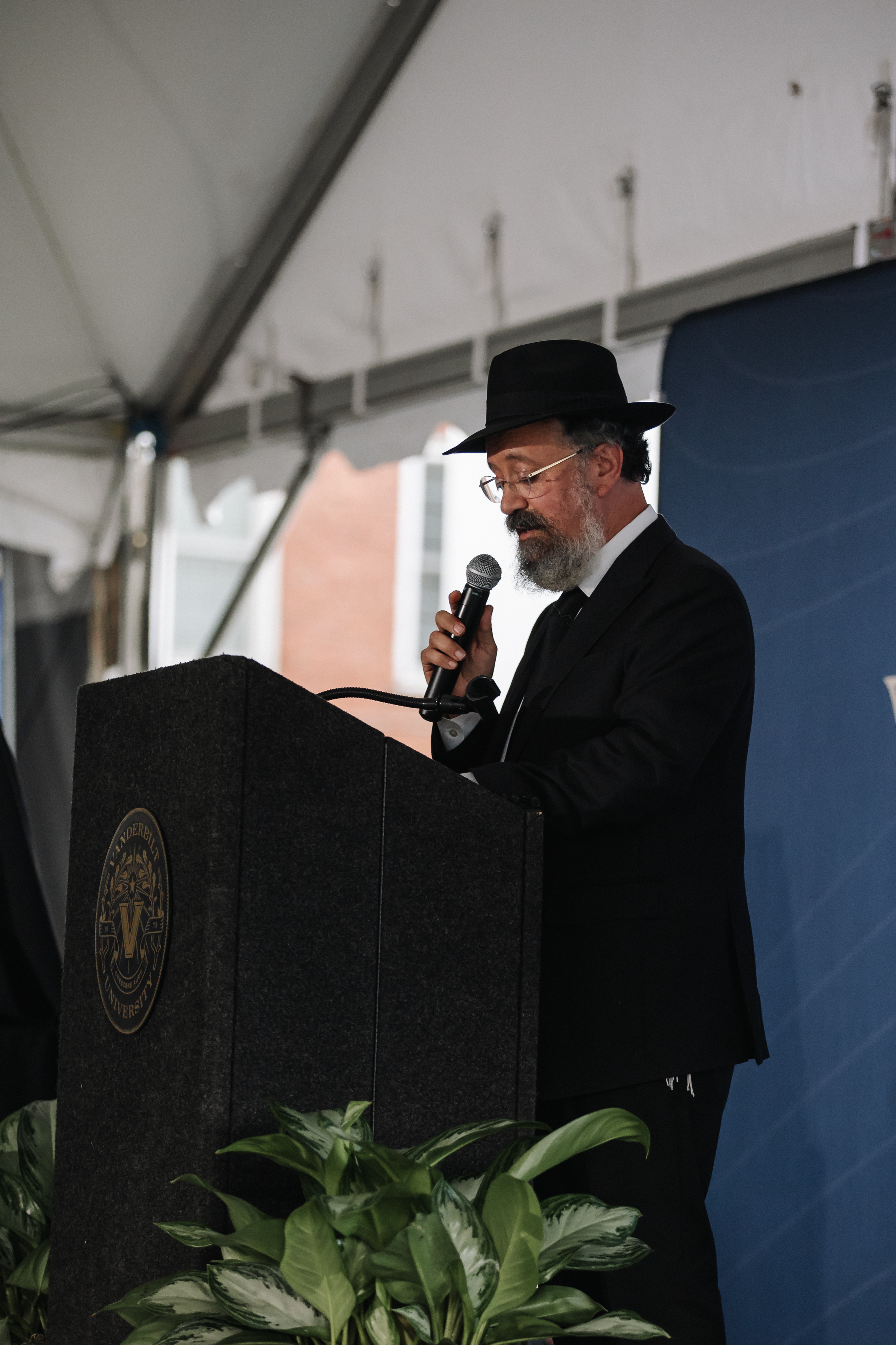 A rabbi in a black hat and suit speaking into a microphone at a lectern during a wedding ceremony.