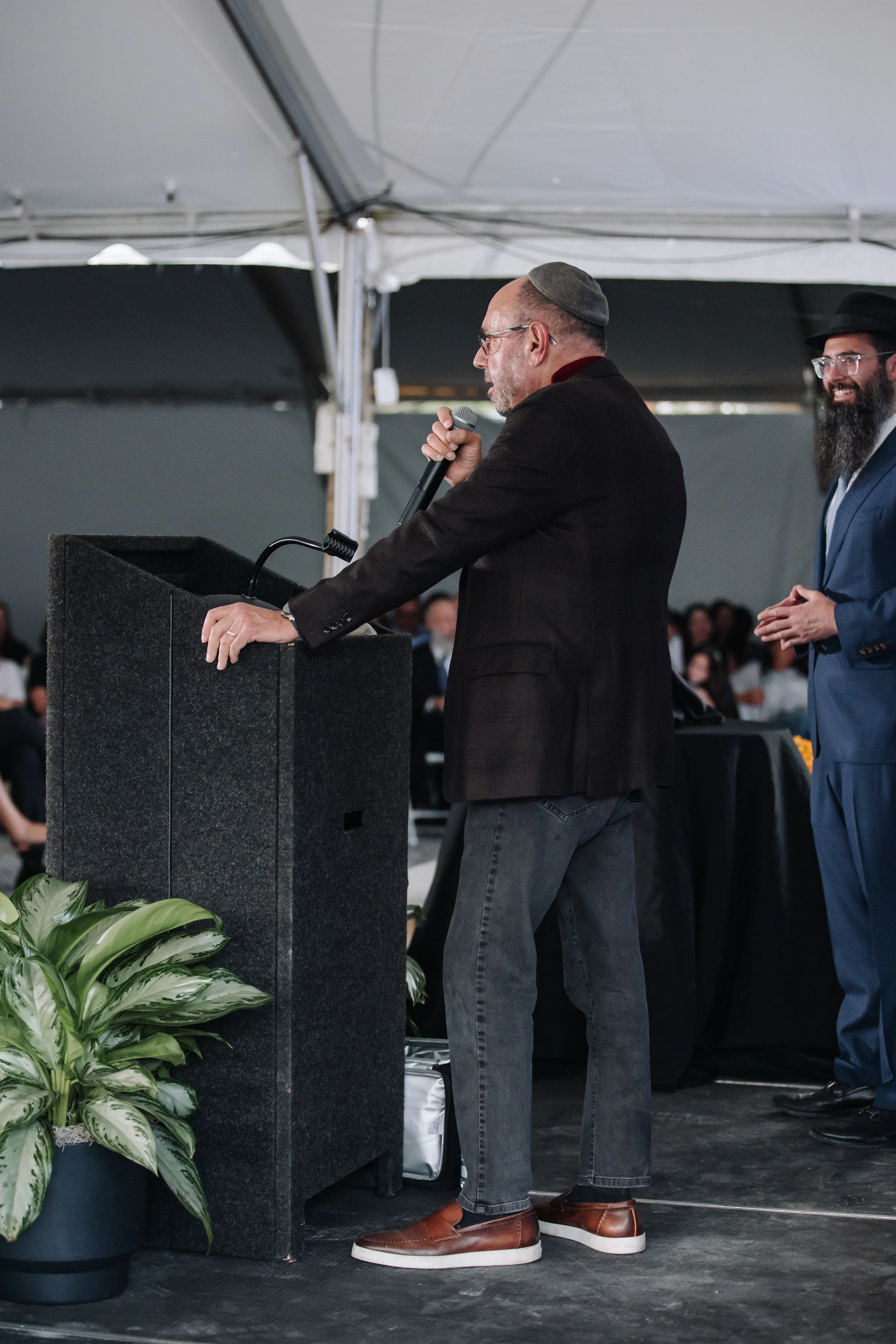 A rabbi speaks into a microphone at a ceremony podium while attendees listen under a tented canopy.