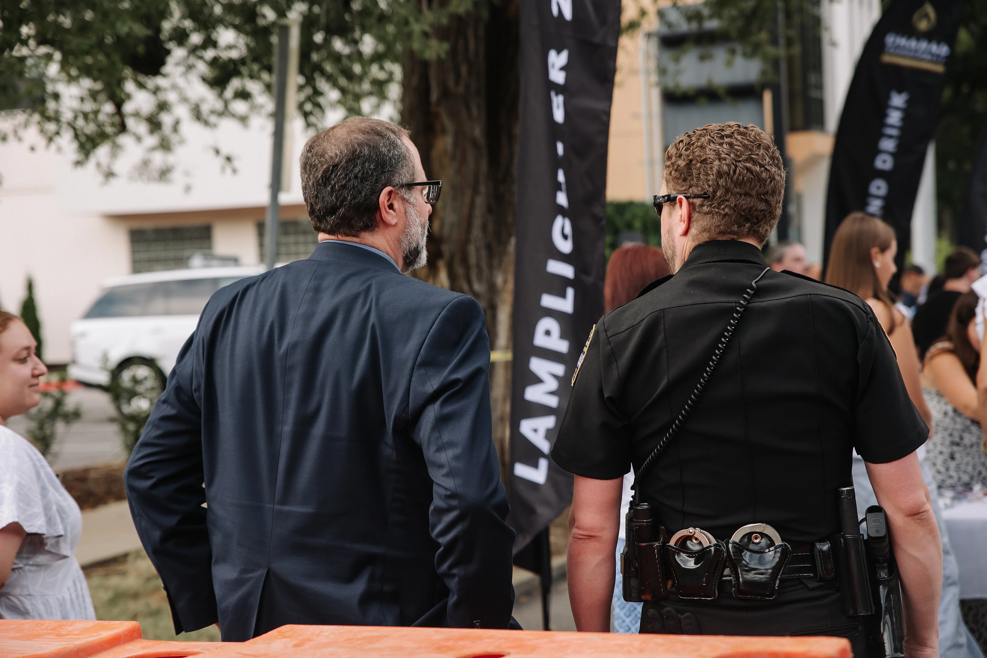 Back view of two men at an outdoor event with a camera operator’s gear visible and event banners in the background.