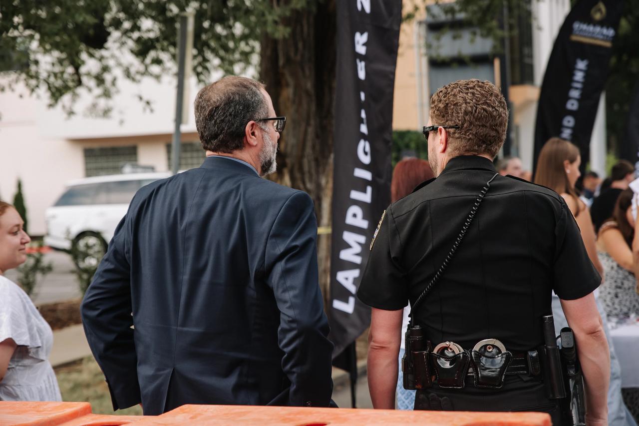 Back view of two men at an outdoor event with a camera operator’s gear visible and event banners in the background.