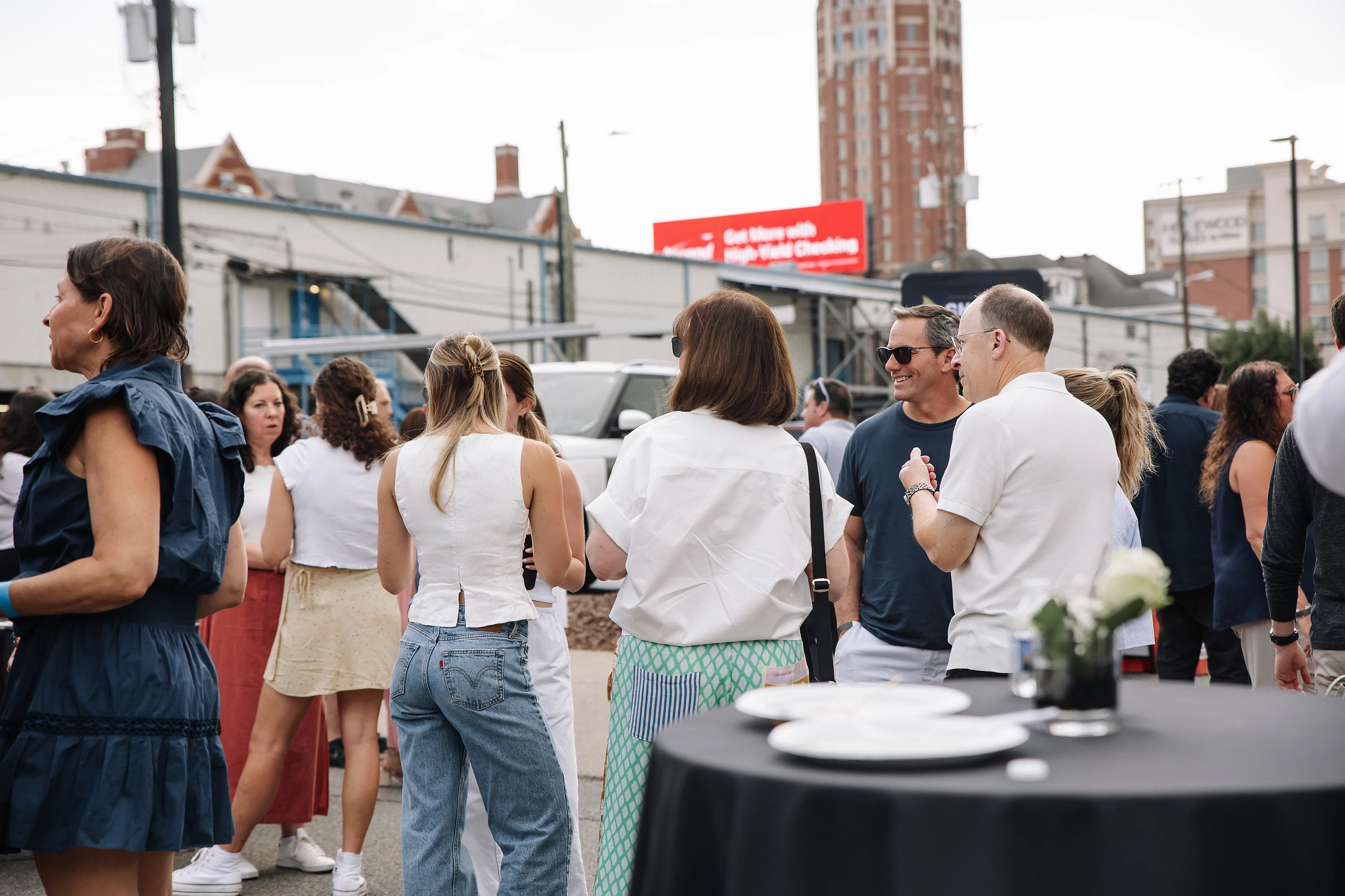 Guests standing and chatting at an outdoor event near a table with plates, with city buildings and tents in the background.