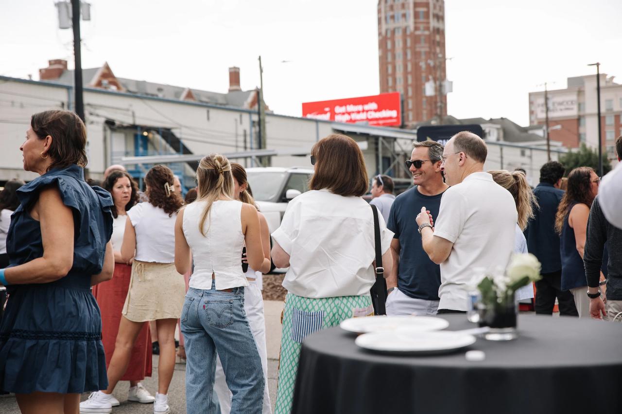 Guests standing and chatting at an outdoor event near a table with plates, with city buildings and tents in the background.