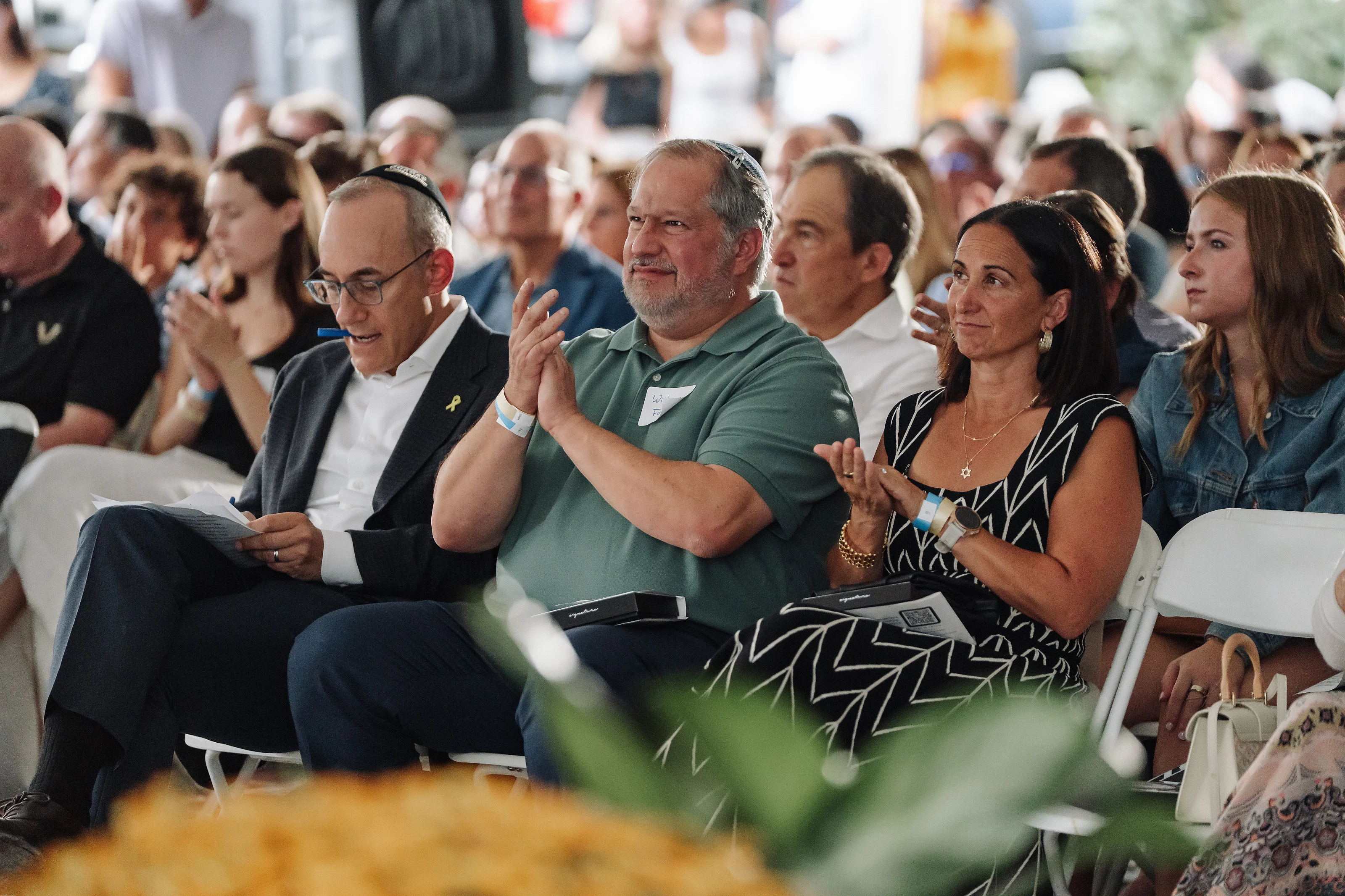 Audience of guests seated at an event, clapping and watching as one man in a green shirt raises his hand.