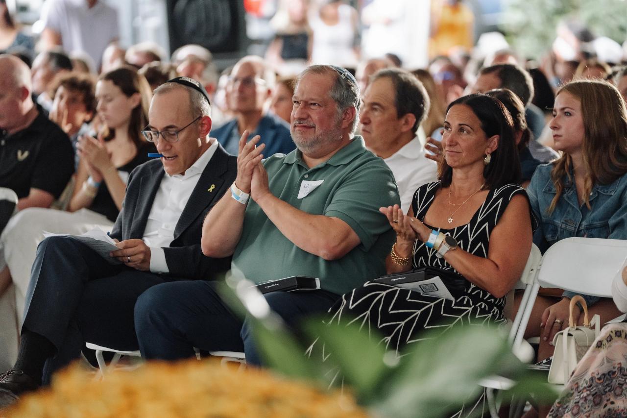 Audience of guests seated at an event, clapping and watching as one man in a green shirt raises his hand.