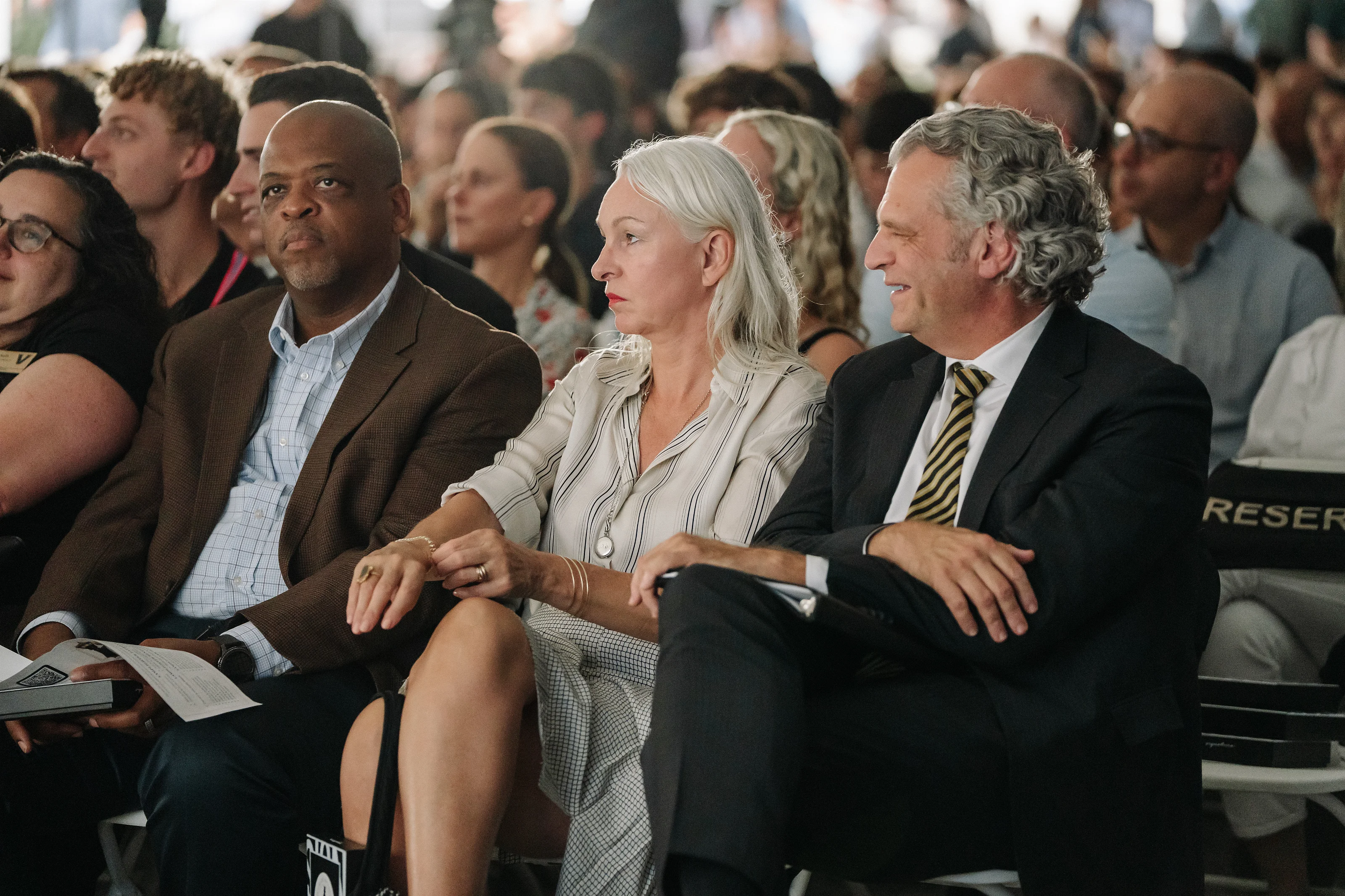 Three seated ceremony guests in the foreground, looking attentively toward the front as the crowd fills the background.