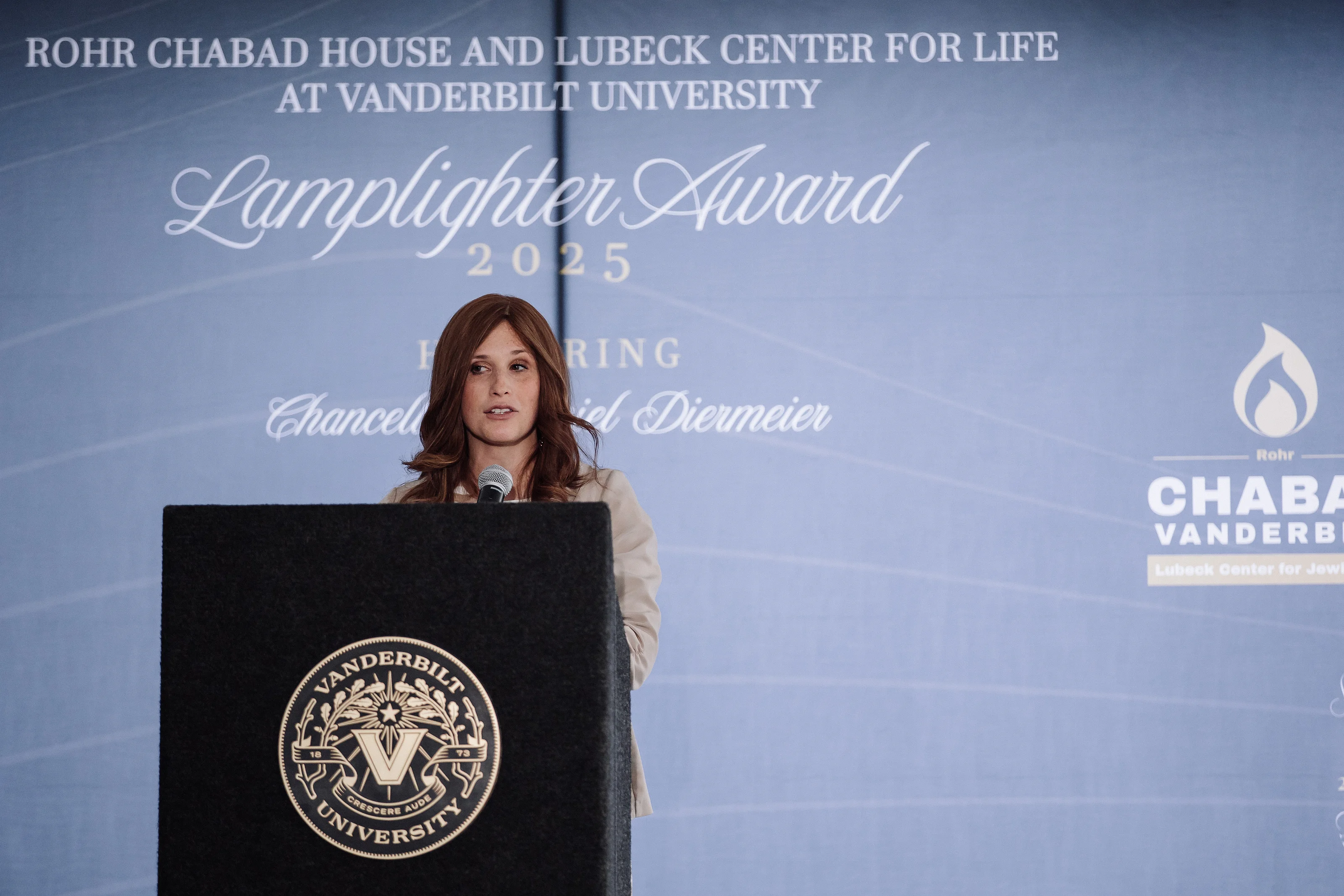 Woman speaking at a podium with microphone in front of a blue event backdrop and award signage