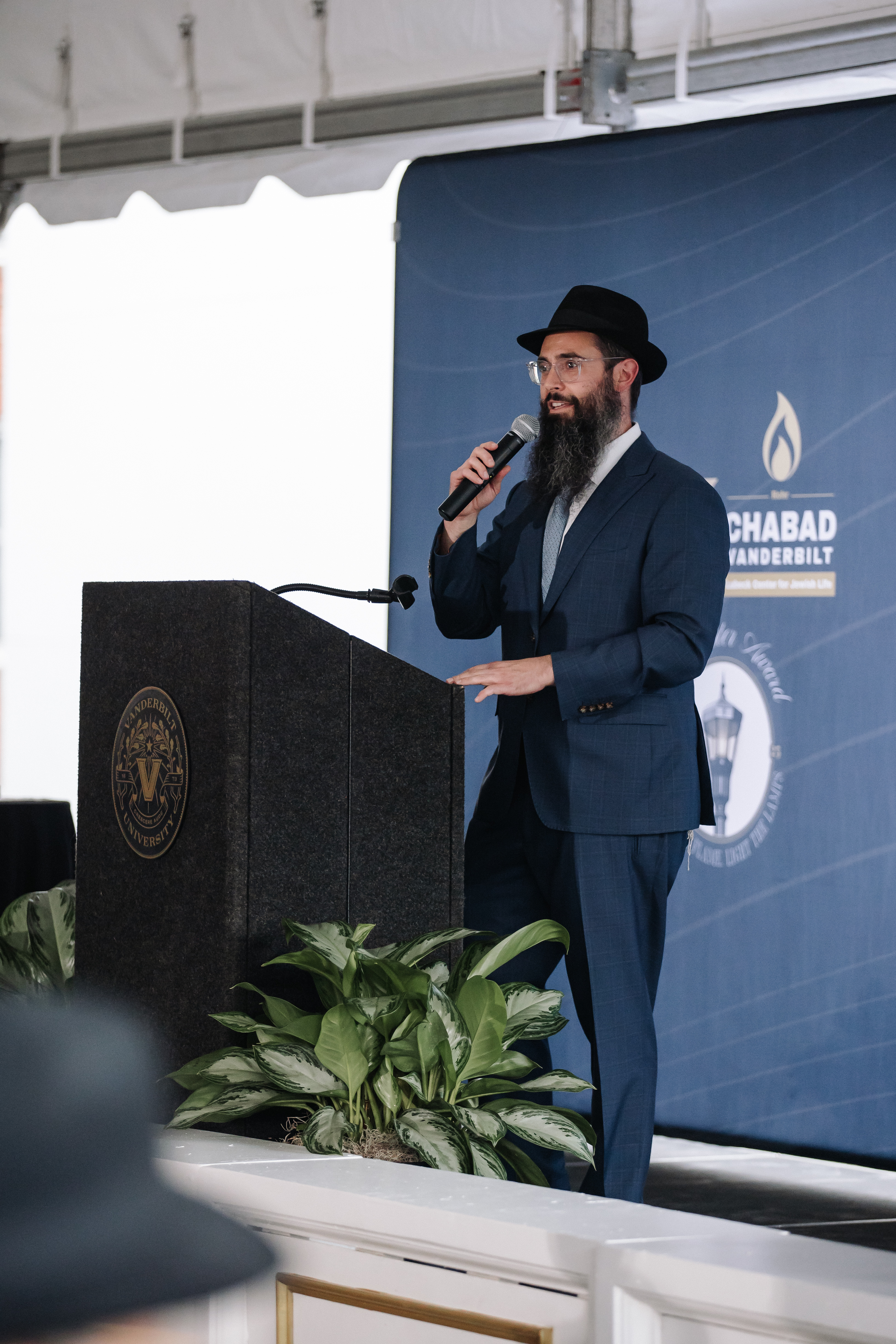 Man in a suit and black hat speaking into a microphone at a lectern against a blue event backdrop.
