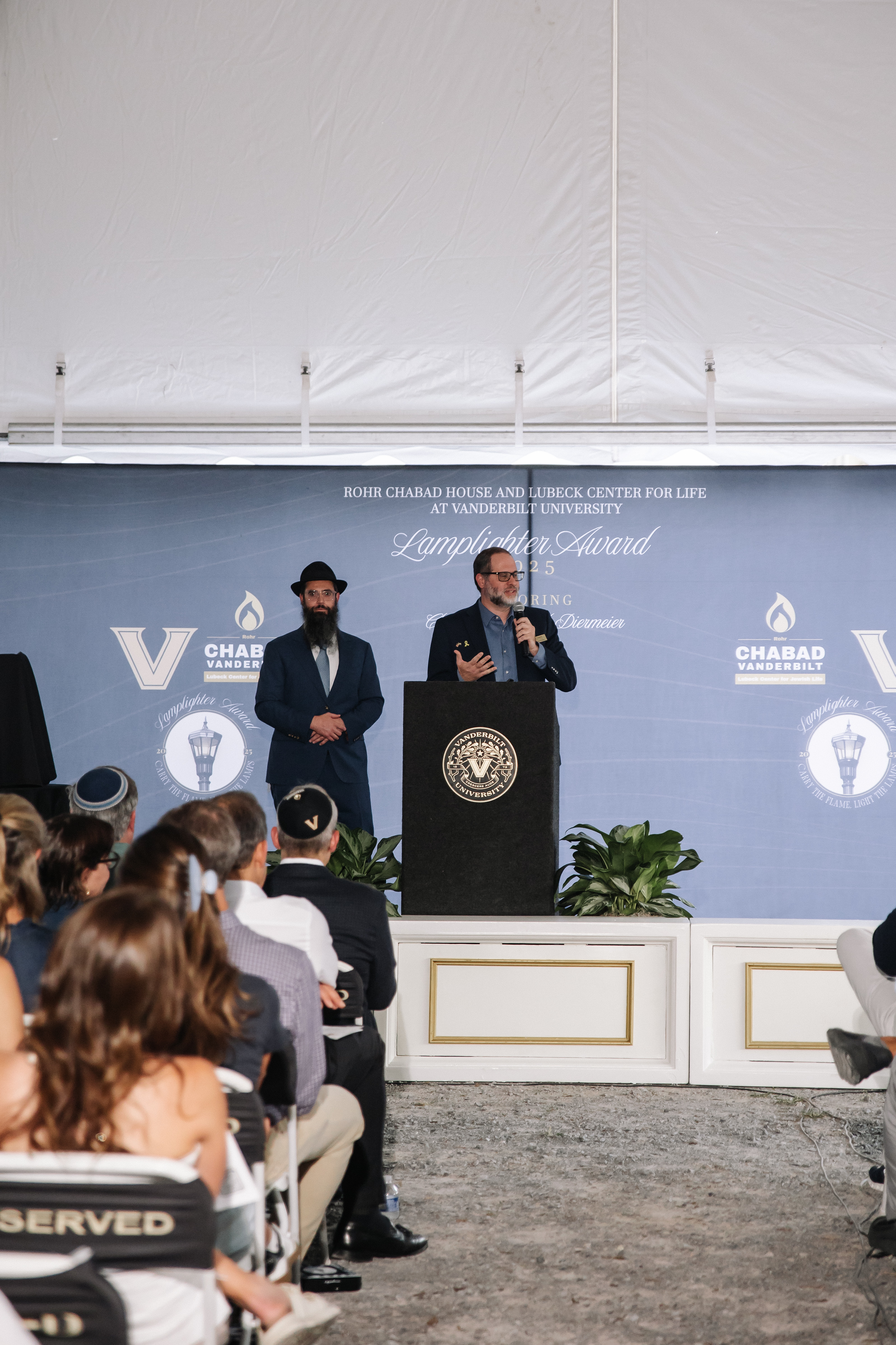 A man speaks at a podium on stage during a community event, with seated attendees in the foreground and event signage on the backdrop.
