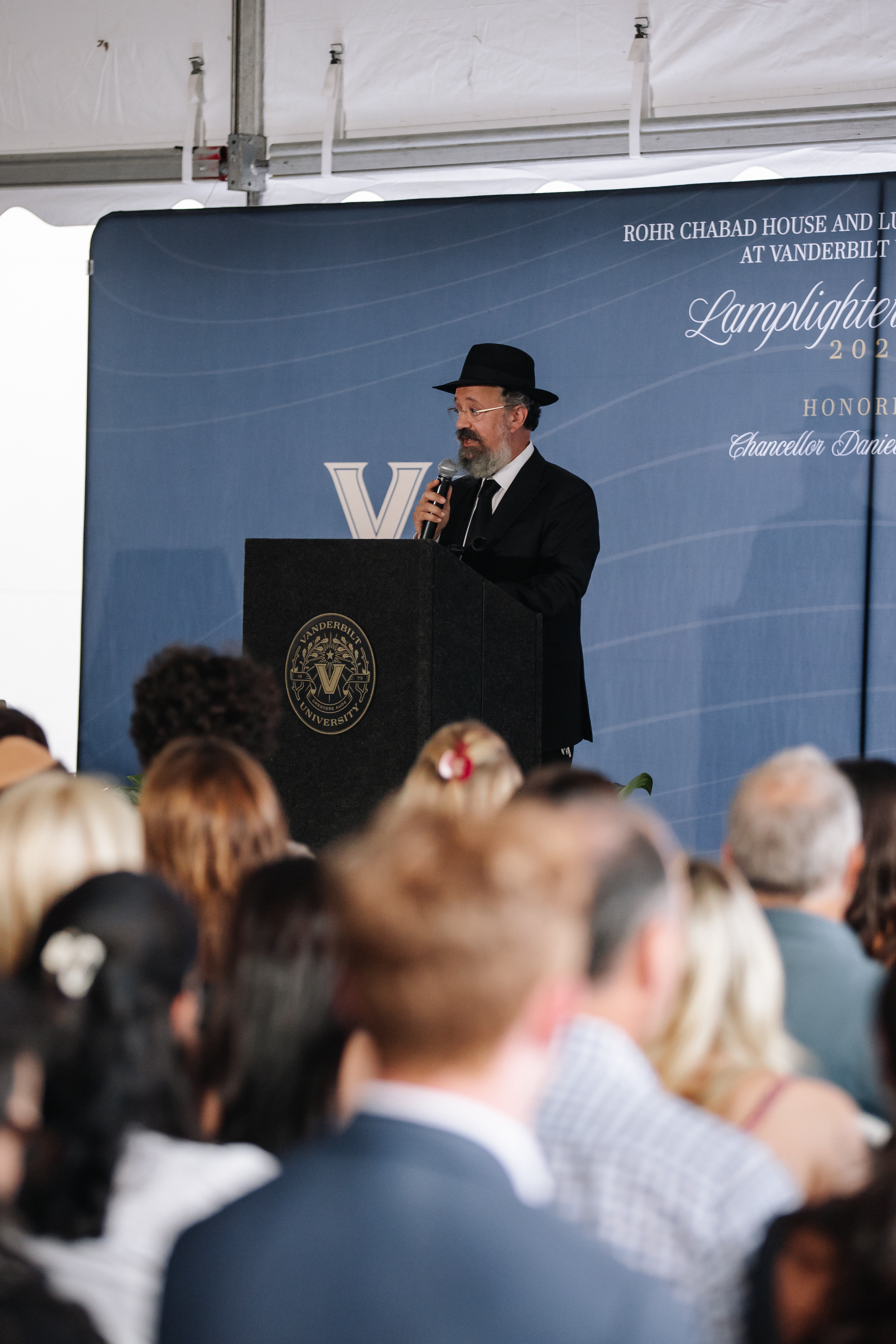 A formal speaker holding a microphone at a podium, addressing a seated audience beneath a blue event banner.