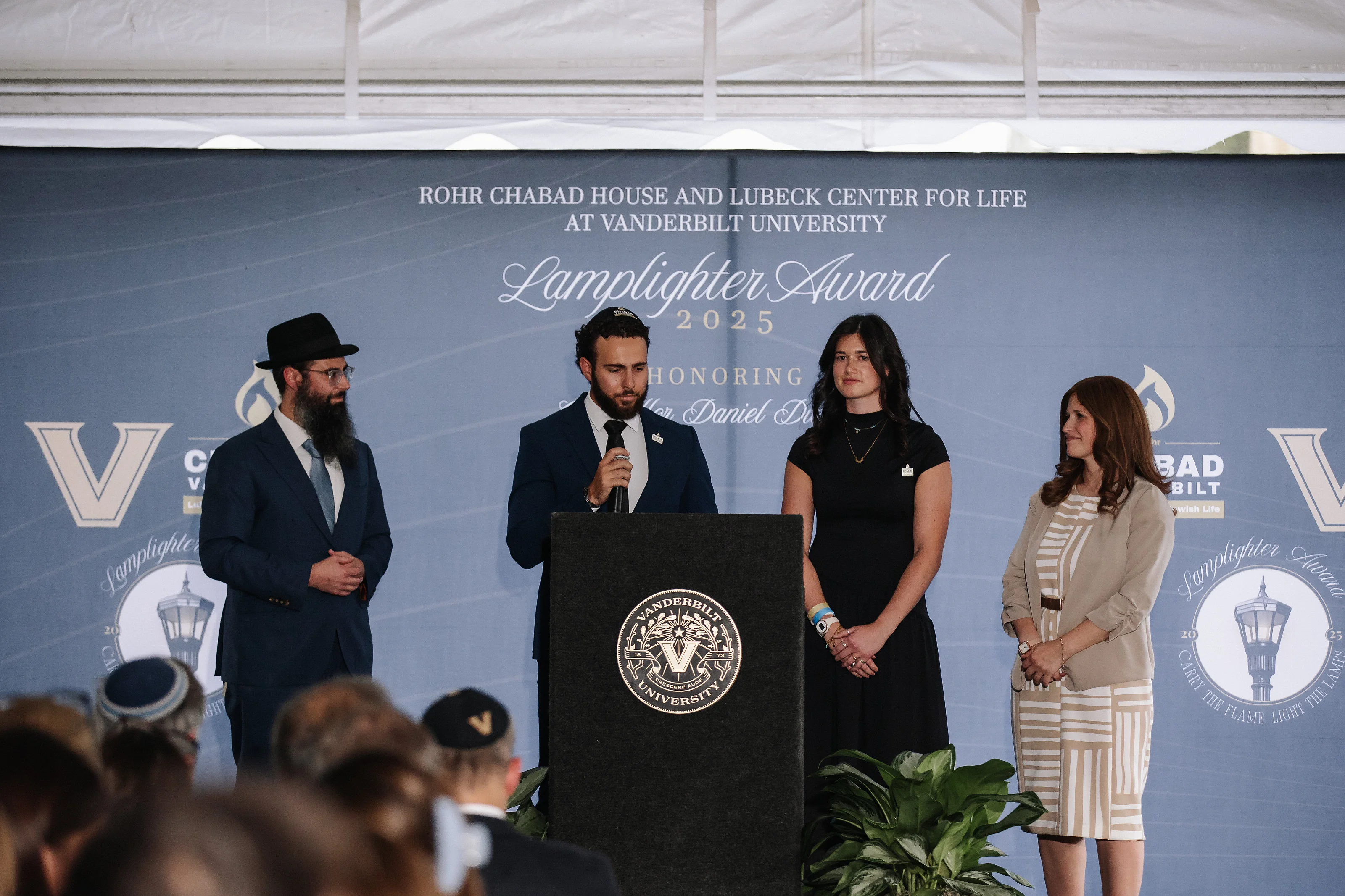 People standing on stage at an award ceremony behind a podium with a university seal, with an audience visible in the foreground.