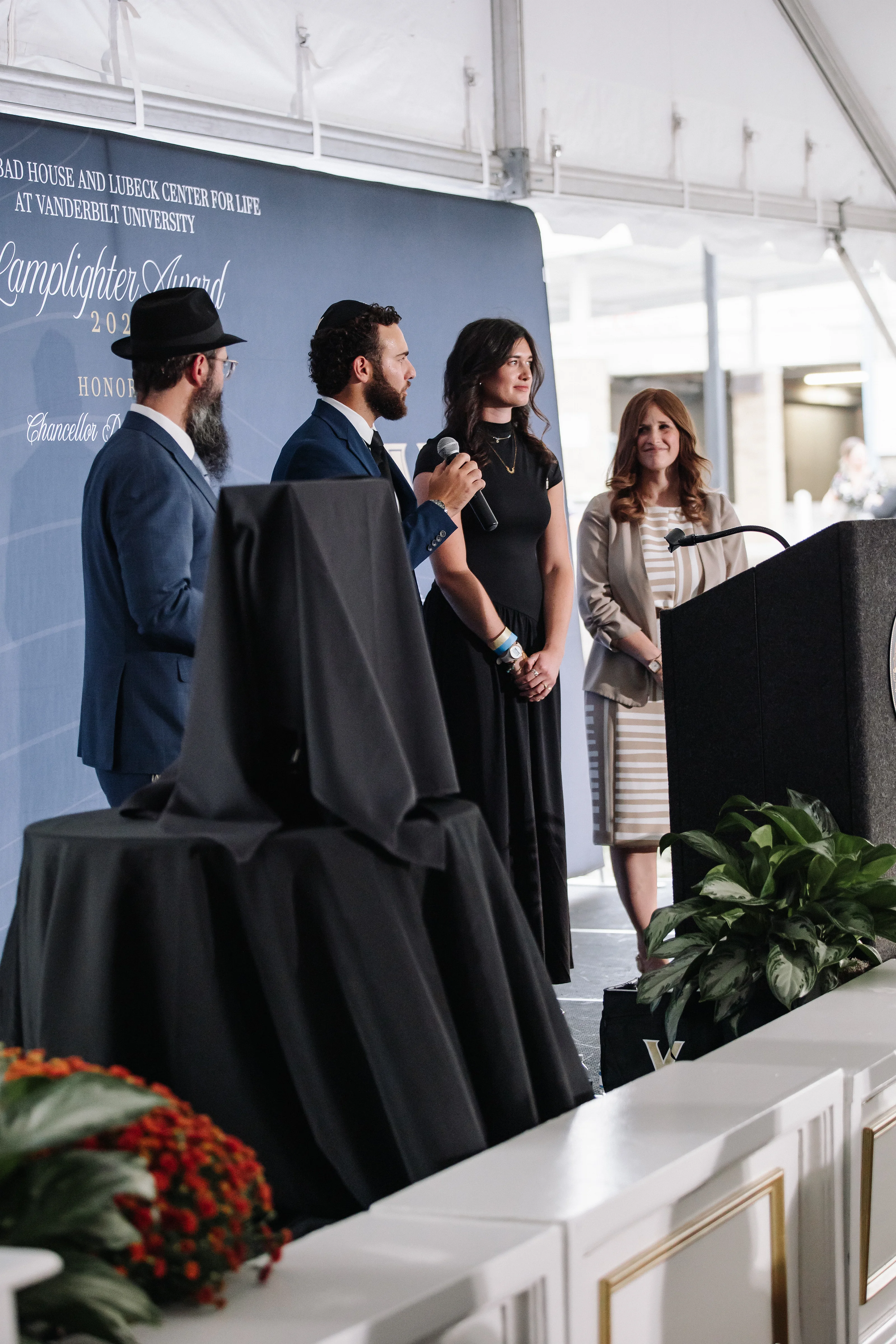 Men and a woman in formal attire stand on a stage near a podium during an event, with one person holding a handheld microphone.