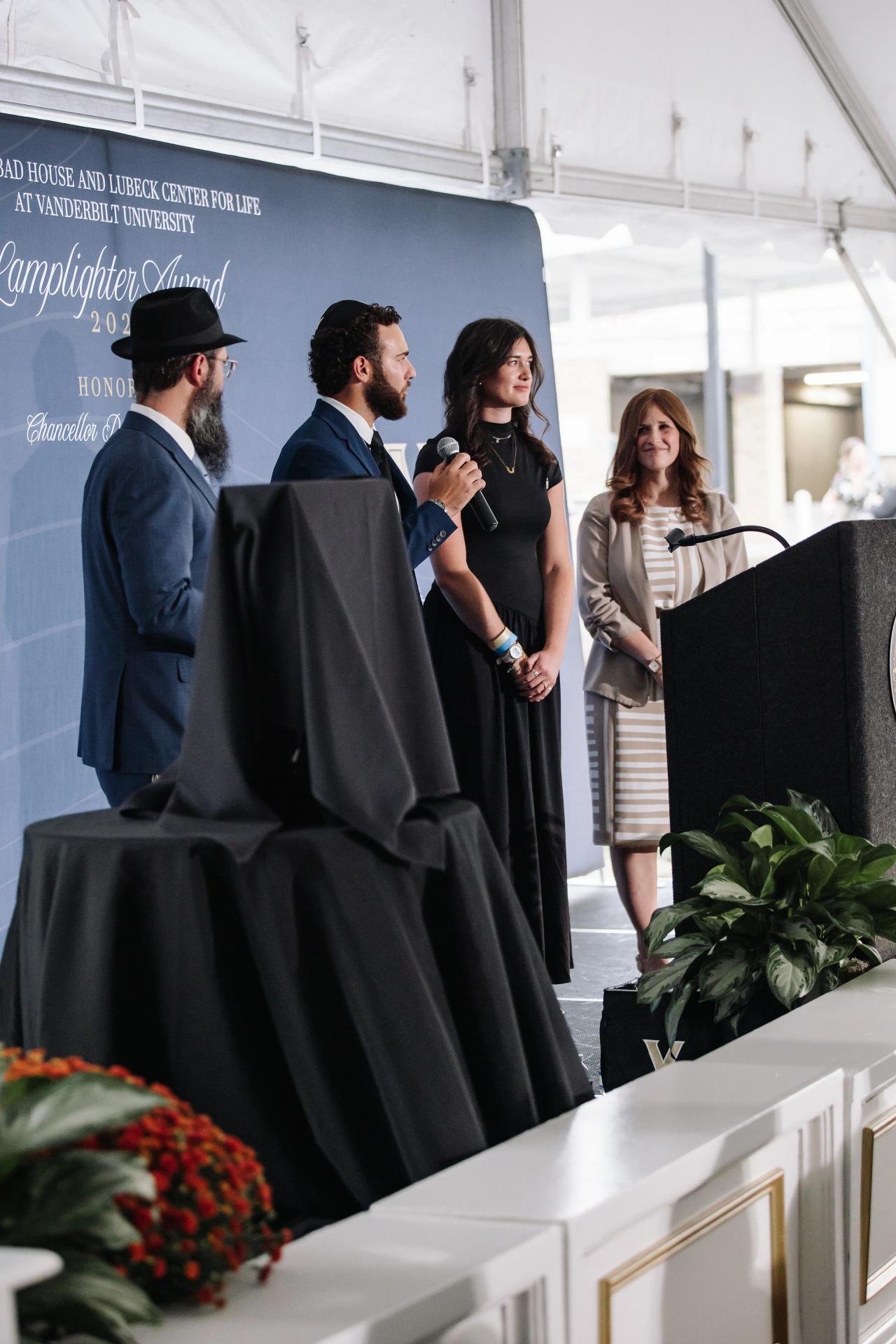 Men and a woman in formal attire stand on a stage near a podium during an event, with one person holding a handheld microphone.