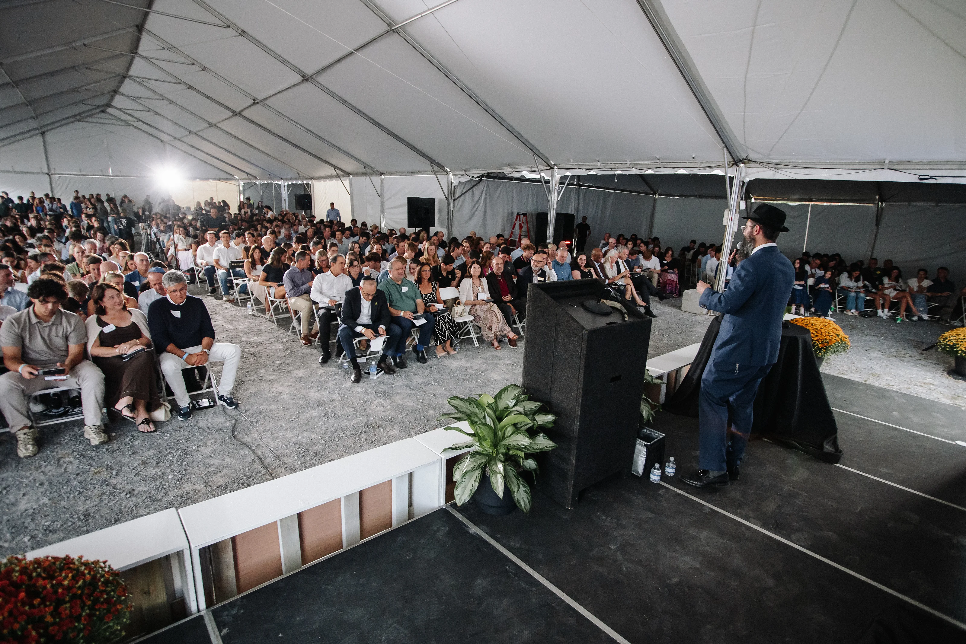 Speaker at a lectern in front of a large seated crowd under a white event tent, during a formal event moment.