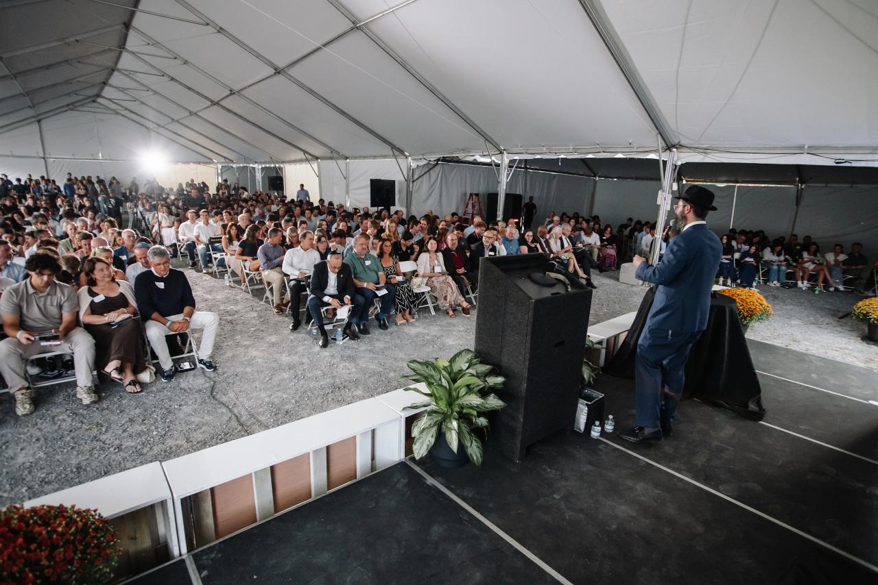 Speaker at a lectern in front of a large seated crowd under a white event tent, during a formal event moment.