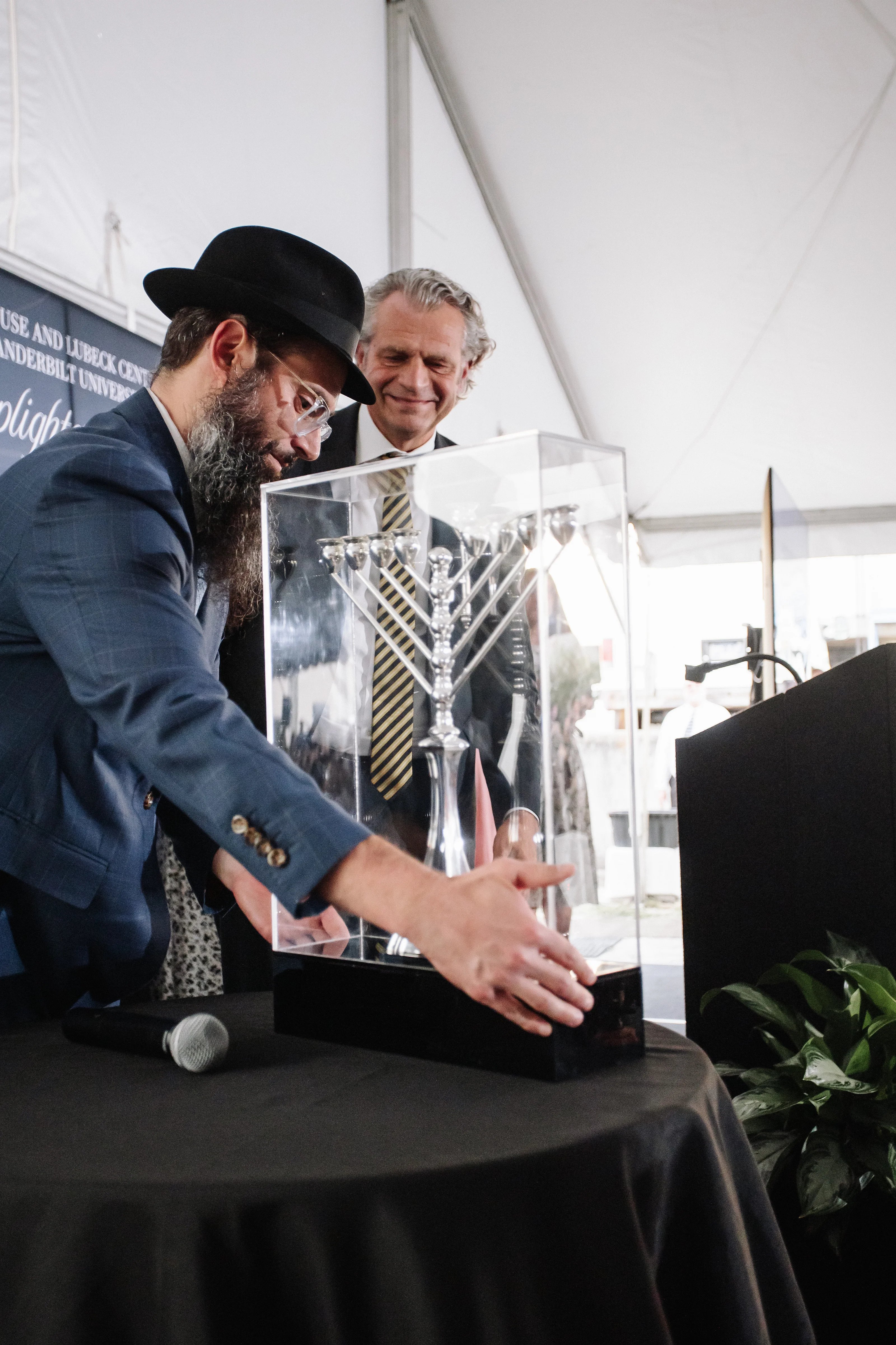 A man in a black hat leans in to sign over a clear acrylic display as another man looks on smiling under a white event tent.