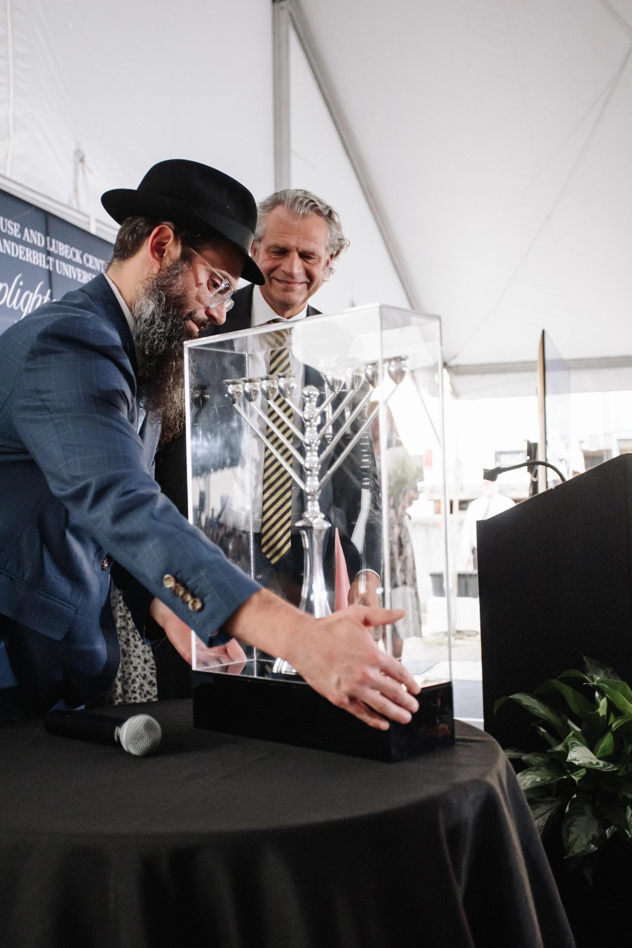 A man in a black hat leans in to sign over a clear acrylic display as another man looks on smiling under a white event tent.