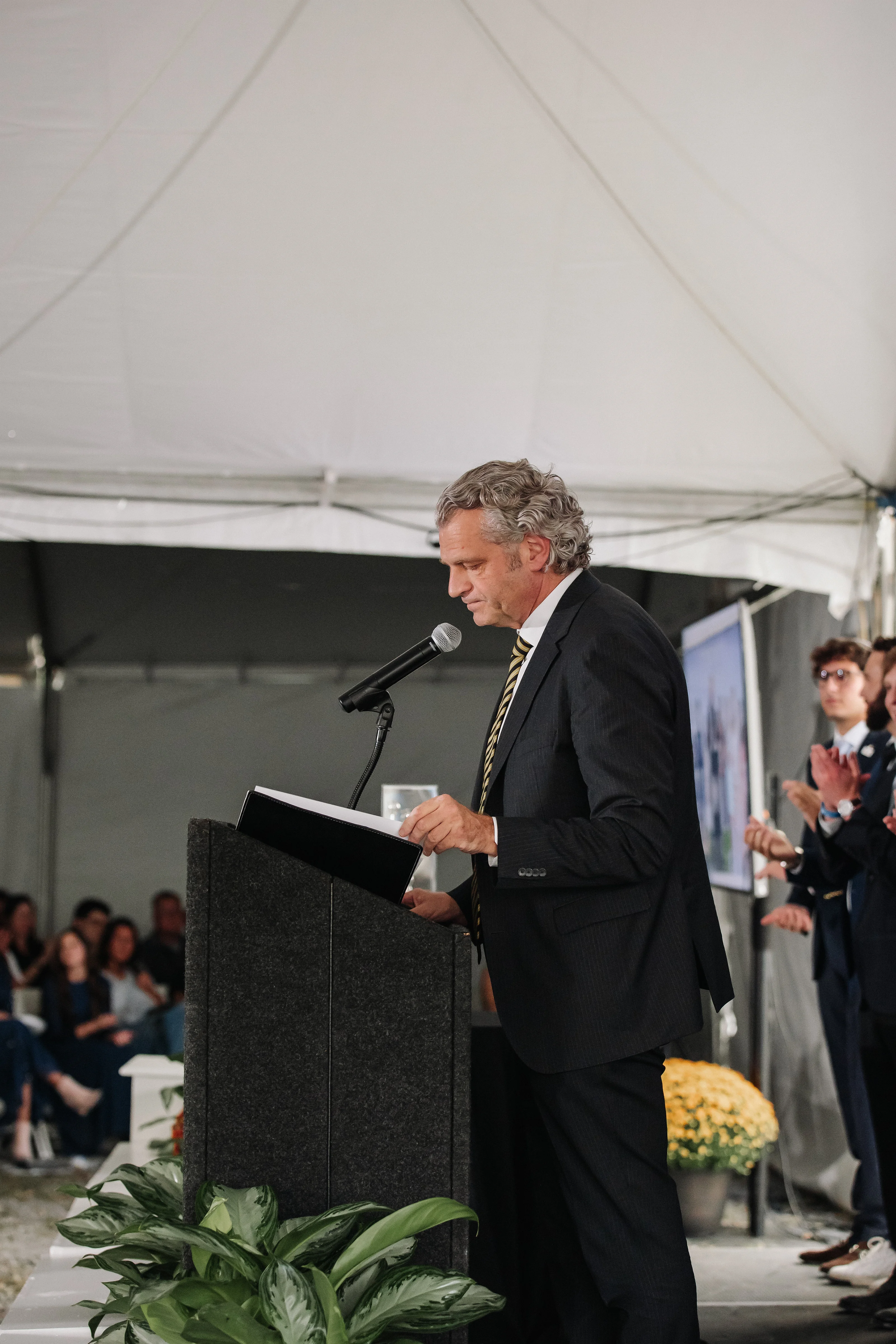 Wedding groom speaking at a podium with microphone under a white tent canopy during a ceremony, with guests seated in the background.