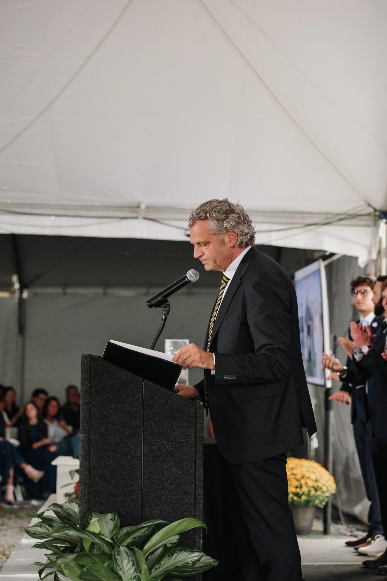 Wedding groom speaking at a podium with microphone under a white tent canopy during a ceremony, with guests seated in the background.