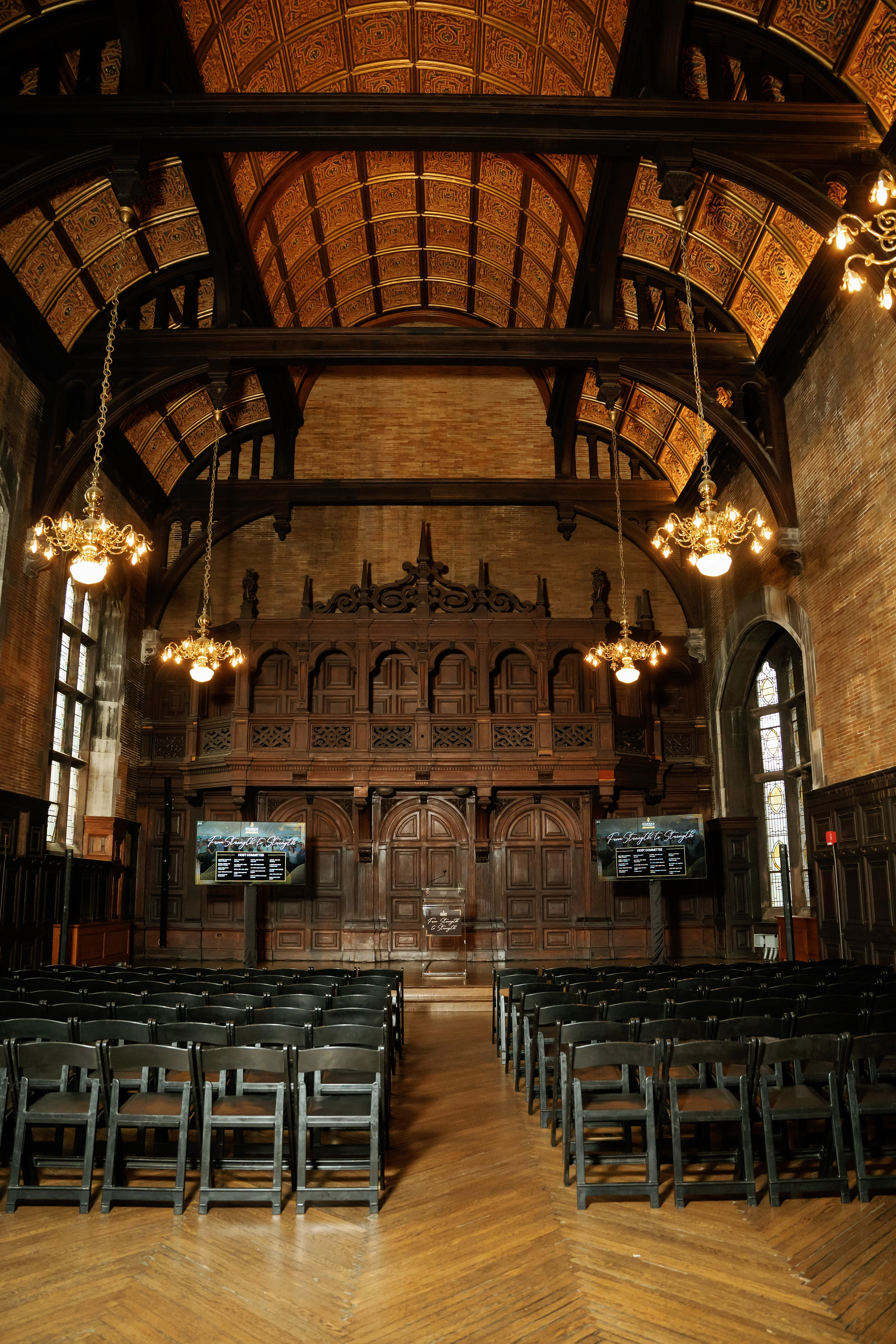 Warmly lit ornate ceremony hall interior with chandeliers, vaulted arches, and empty chairs facing the stage.