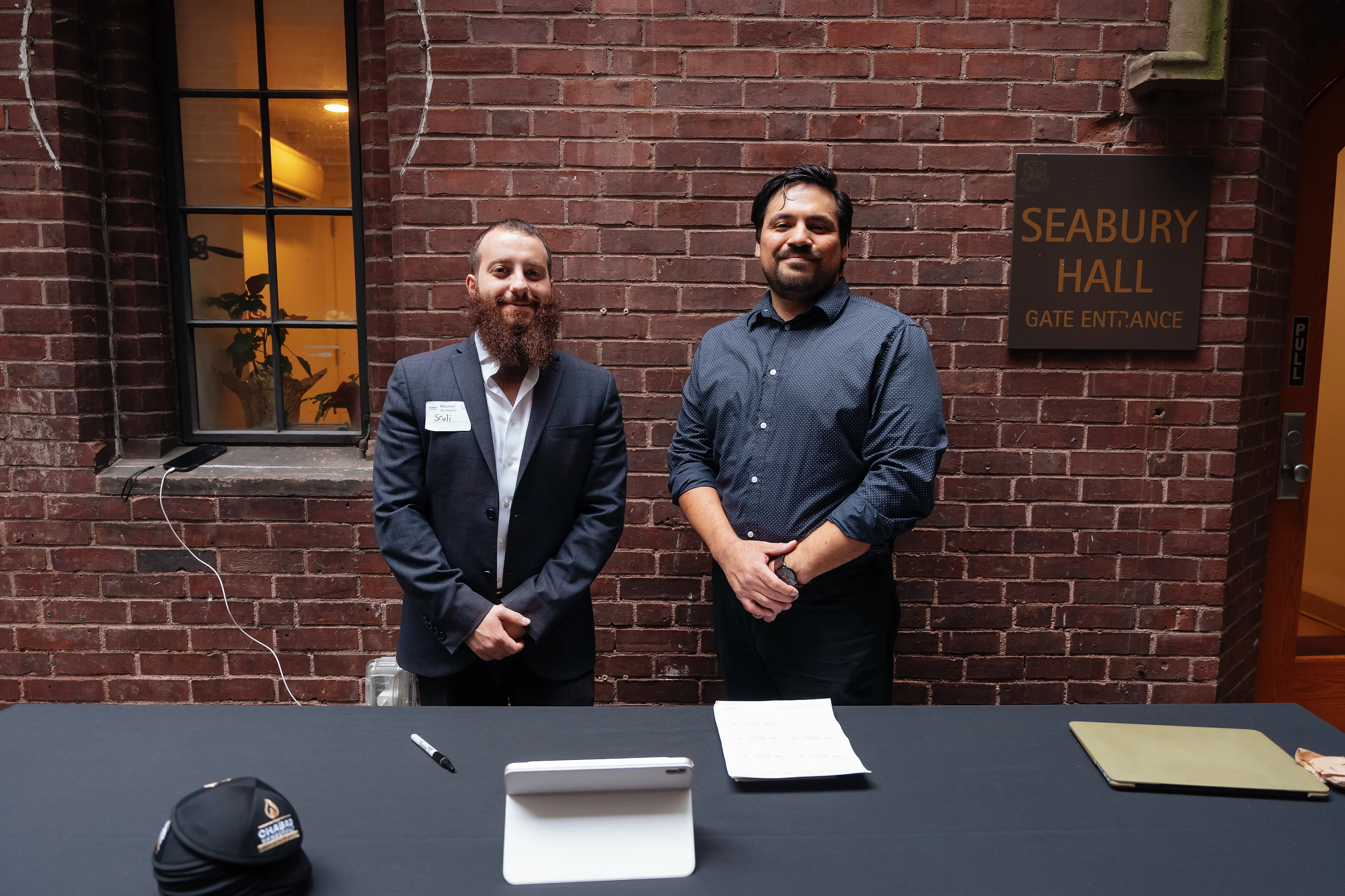 Two men standing behind an event check-in table in front of a brick wall, with event signage in the background.