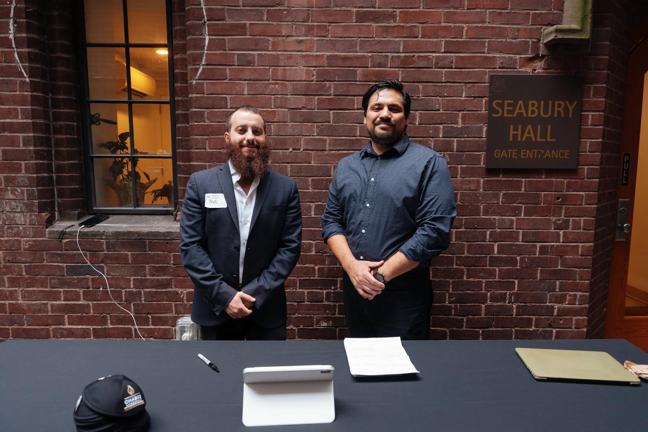 Two men standing behind an event check-in table in front of a brick wall, with event signage in the background.