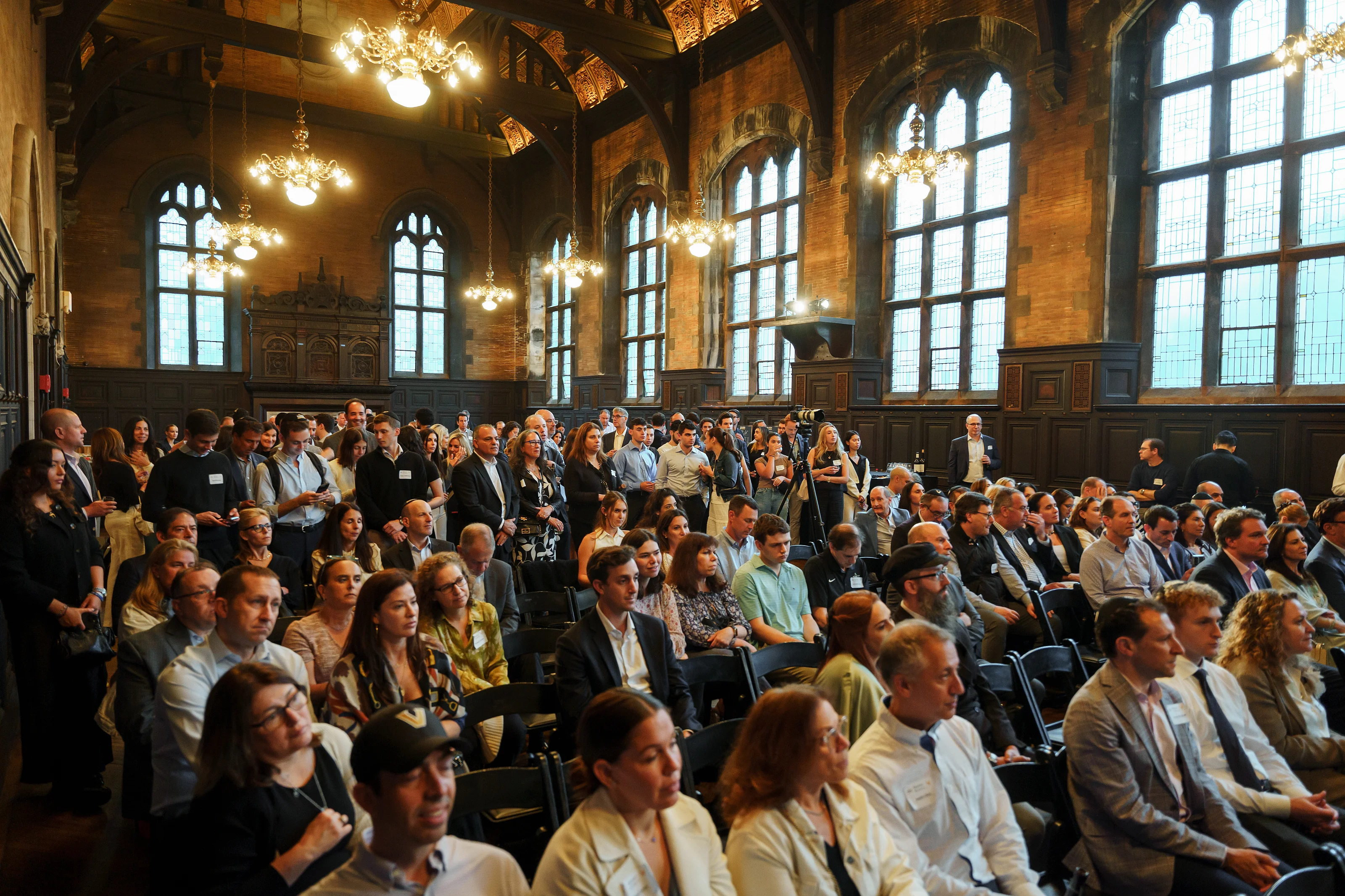 Wedding/mitzvah ceremony audience seated in a grand hall with tall arched windows and warm chandelier light.