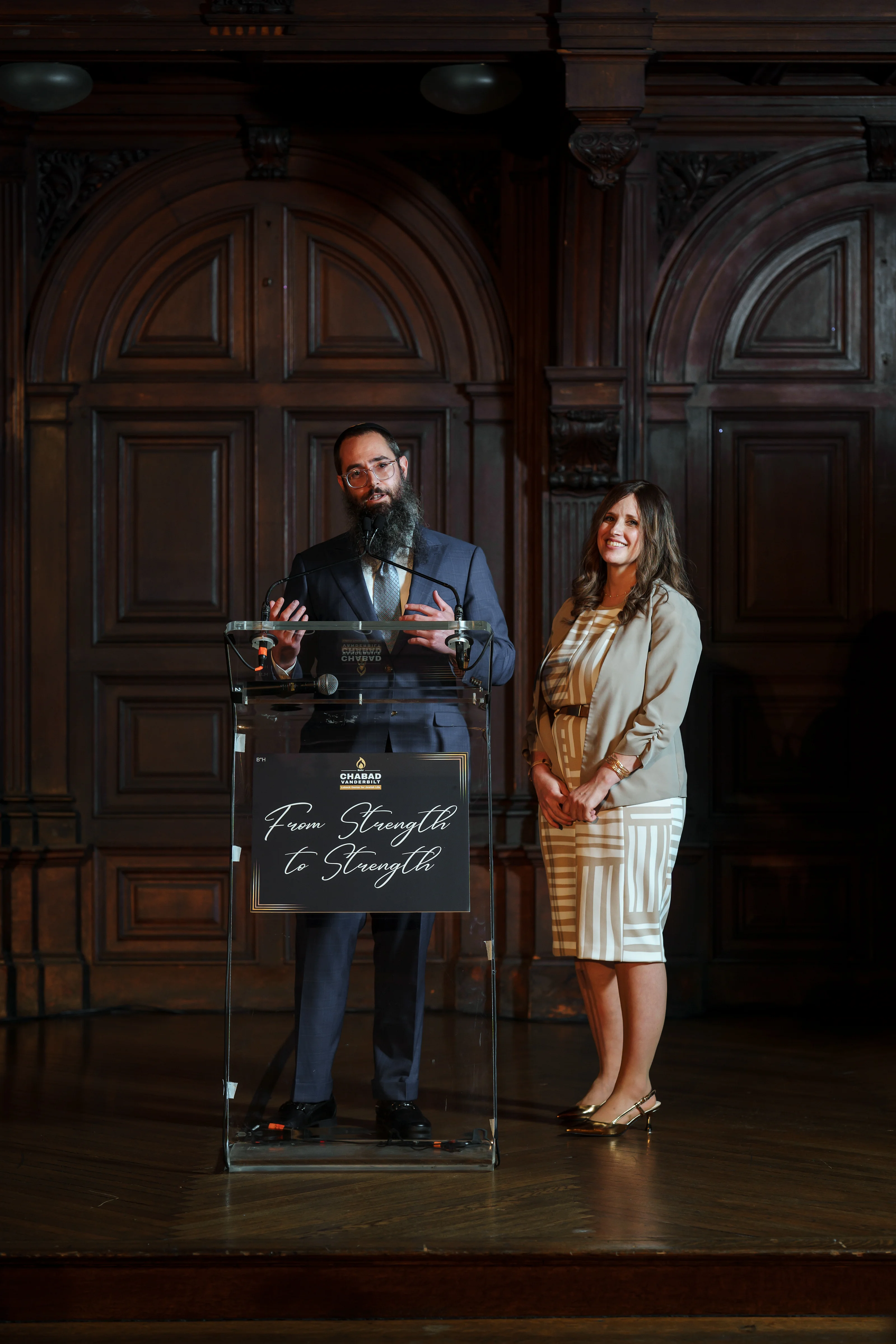 Couple standing near a clear ceremonial podium on a stage with dark wood paneling, lit by warm overhead lighting.