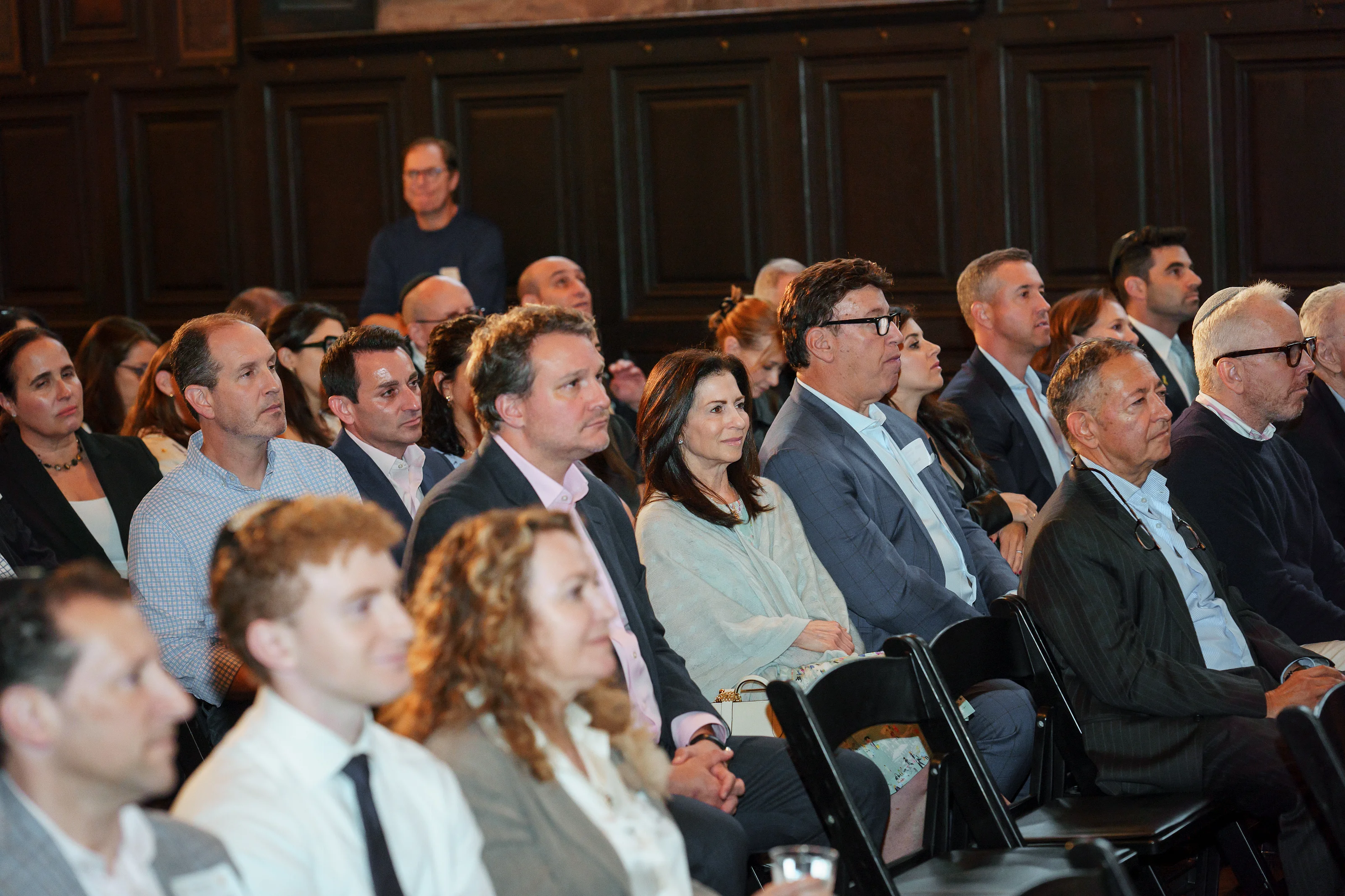Guests seated in a ceremony hall, viewed from behind a shallow depth-of-field foreground row and facing forward under warm indoor light.
