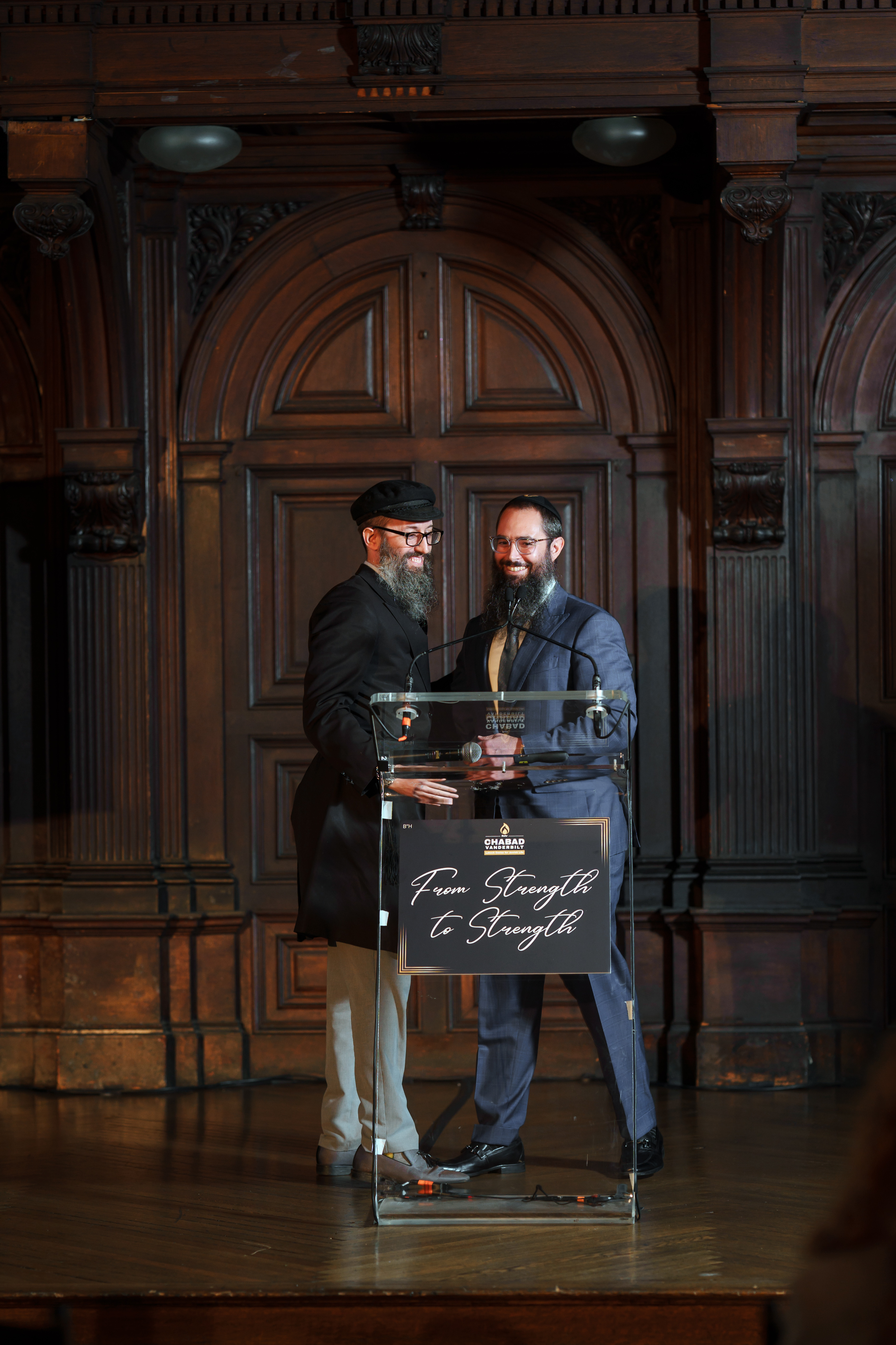 Two bearded men in formal black attire stand behind a clear lectern on a stage with an ornate arched wooden backdrop, smiling during an event presentation.