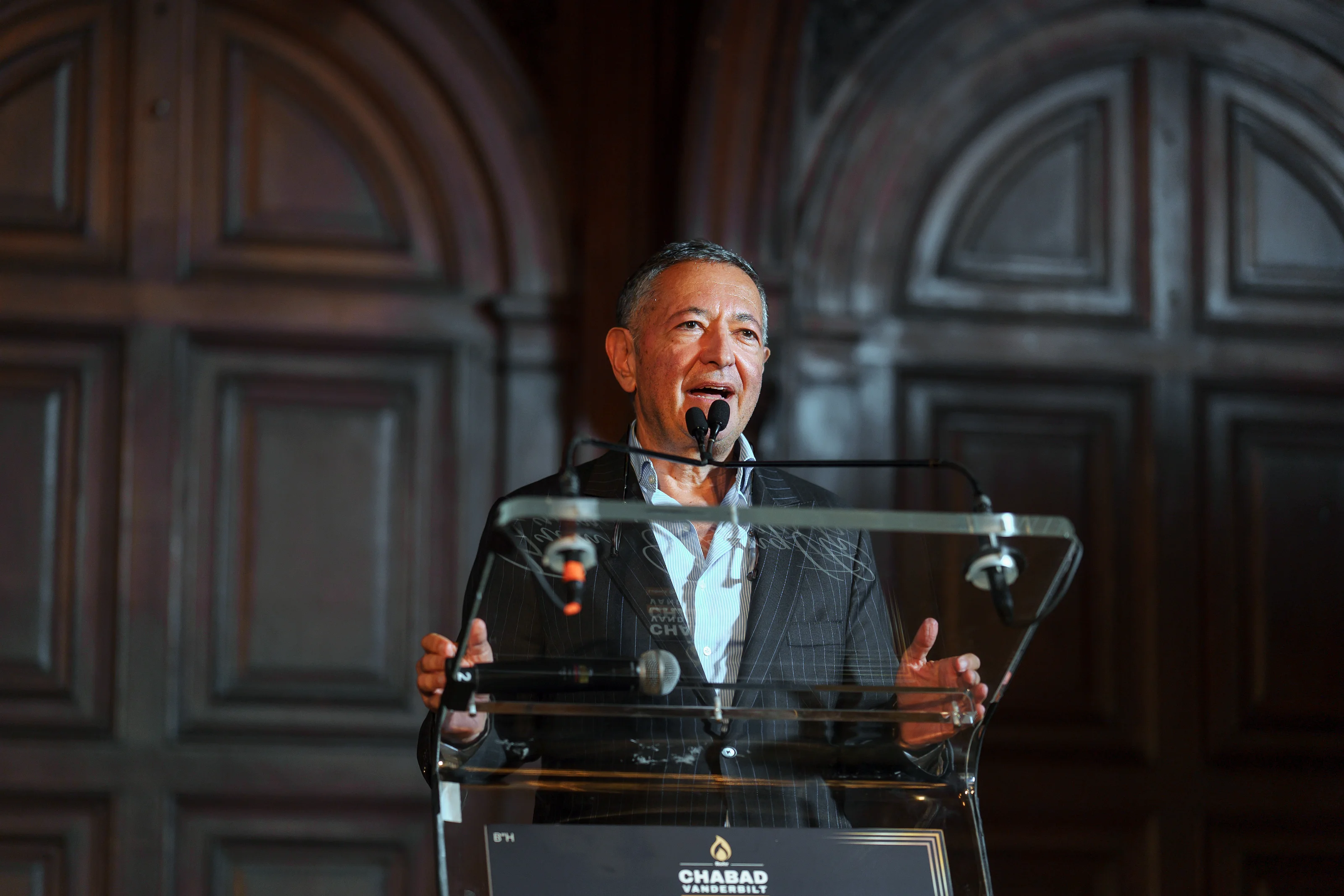 Framed speaker at a microphone behind a clear podium in an indoor ballroom with arched architectural backdrop