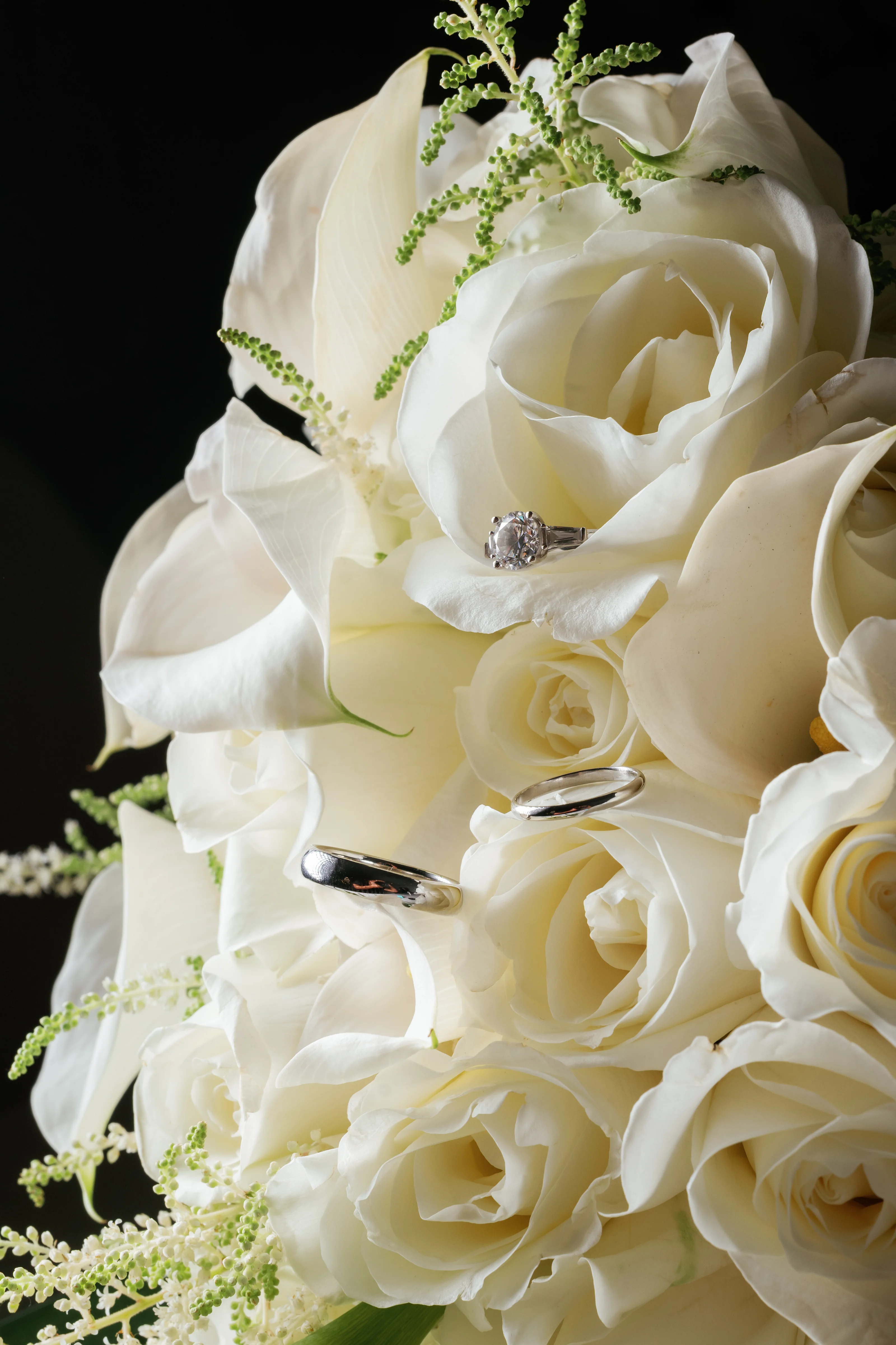 White wedding rings resting on a bouquet of white roses with green greenery, shot against a dark background.